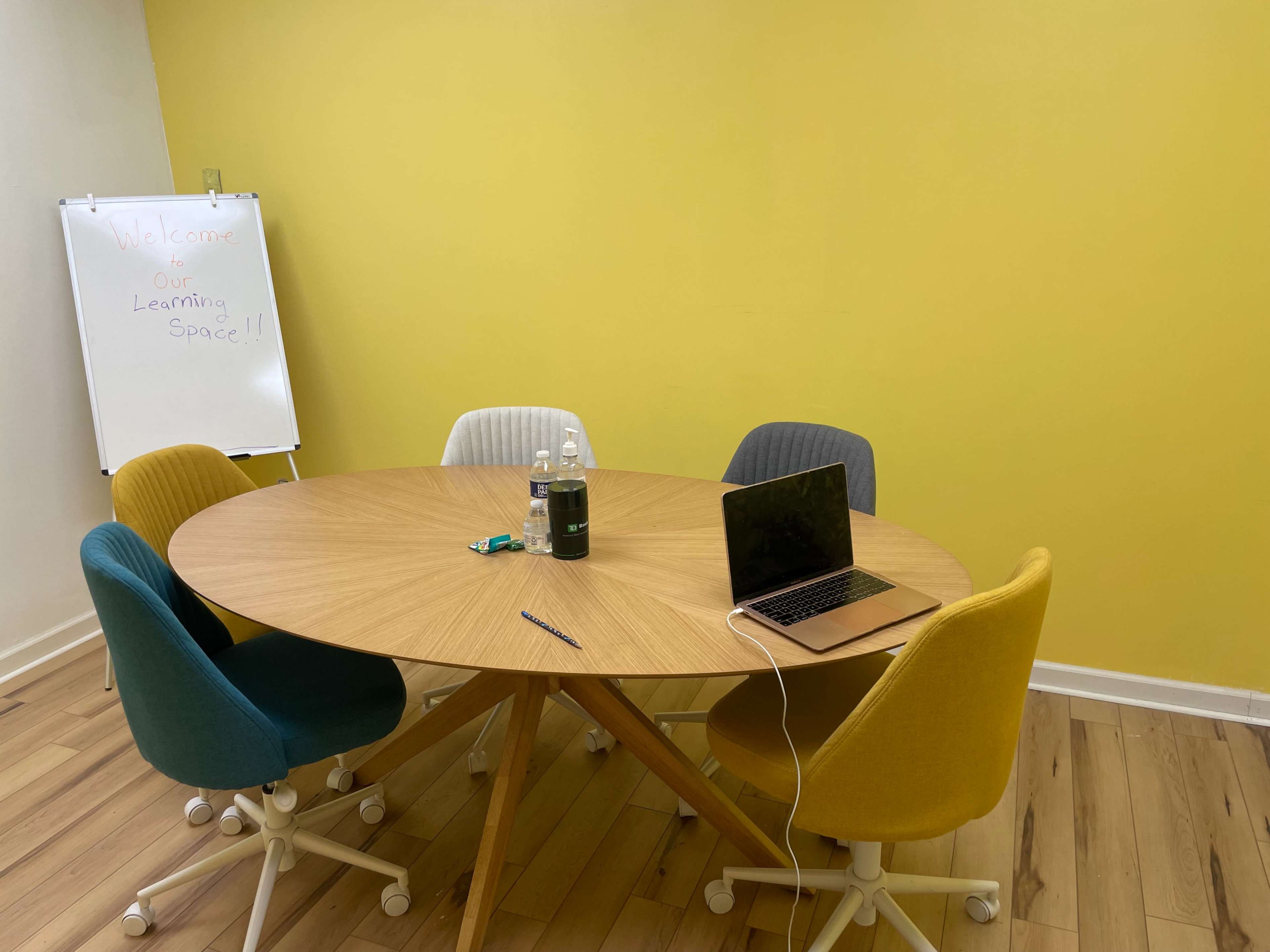 A round wooden table surrounded by five chairs in a room with a yellow wall and a whiteboard.