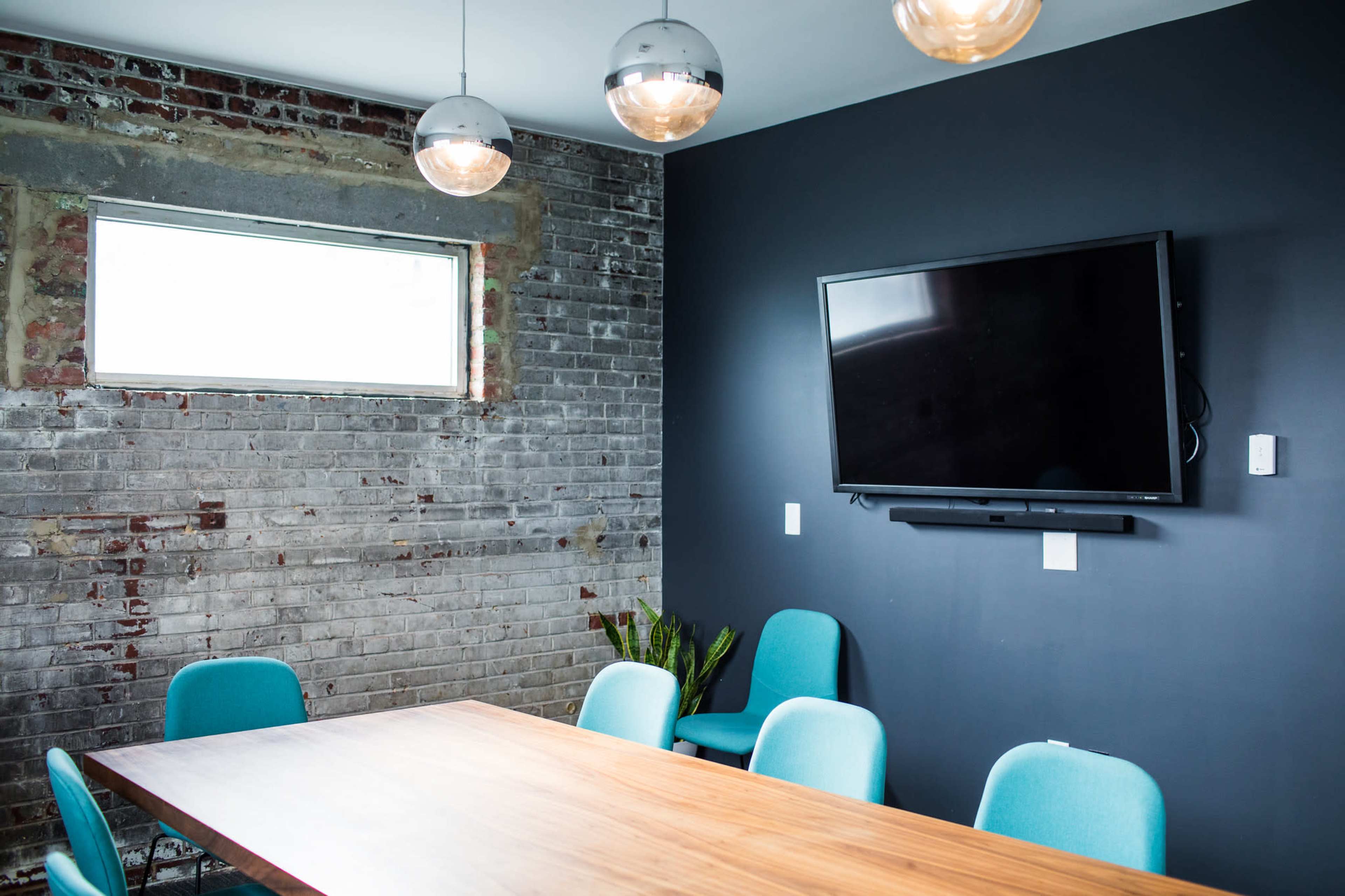 A conference room features a wooden table surrounded by blue chairs, with a flat-screen TV mounted on a dark wall and a window providing natural light.