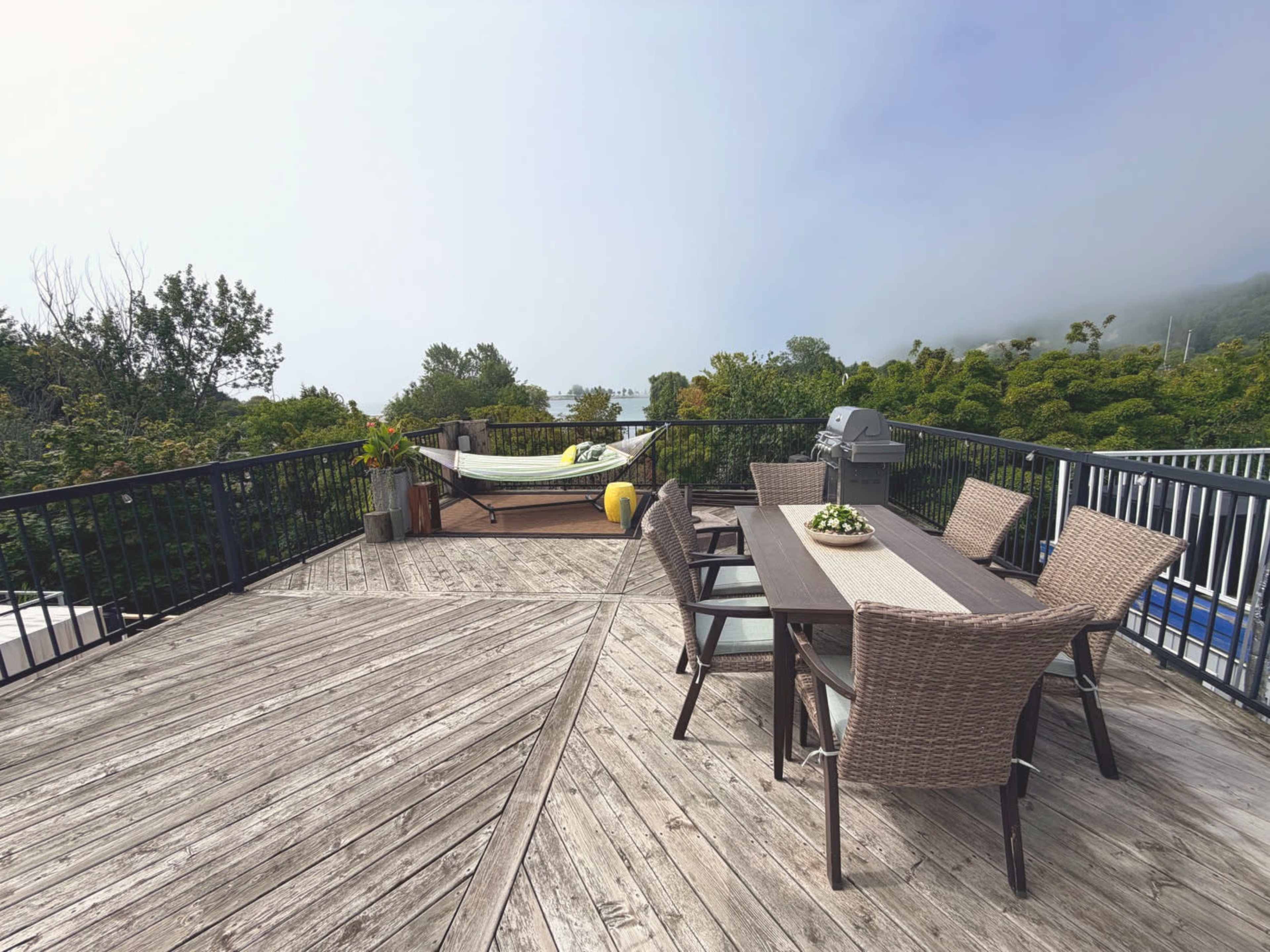 A spacious wooden deck features a dining table with chairs, a hammock in the background, and a barbecue grill amidst greenery and fog.