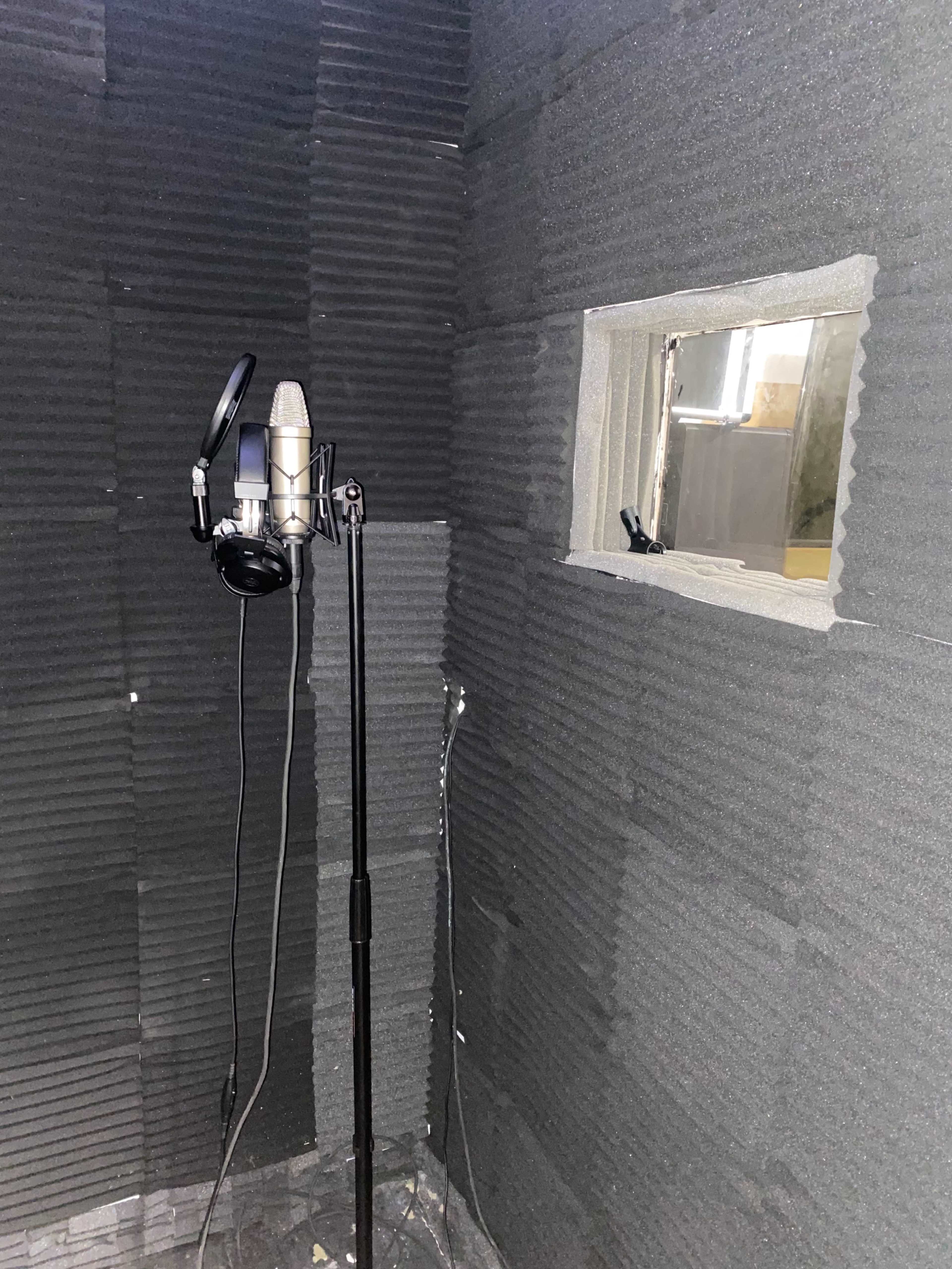 The image shows a soundproof recording booth featuring a microphone on a stand and a small window.