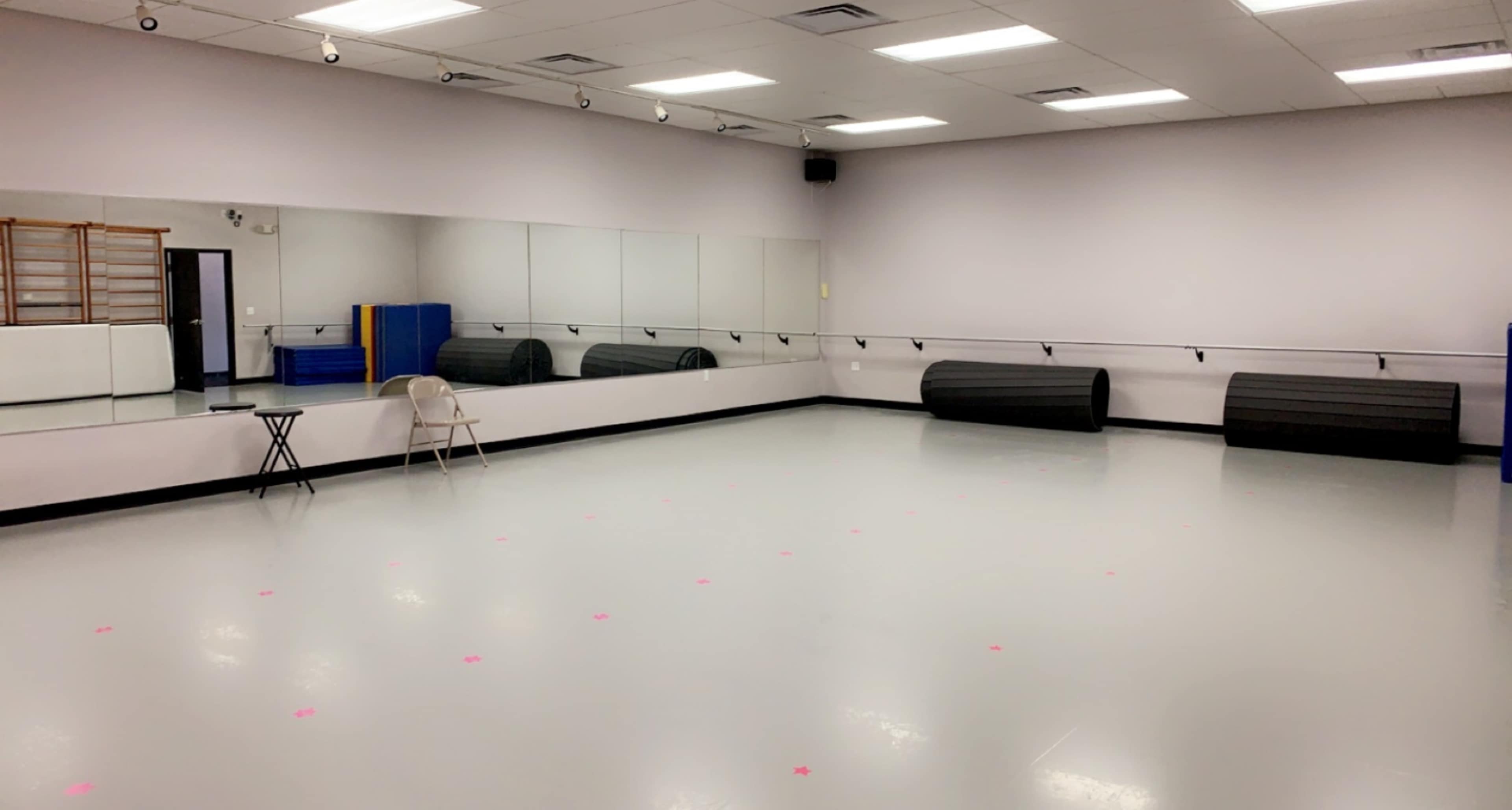 A spacious dance studio with mirrored walls, a gray floor, and two black mats rolled up against the back wall.