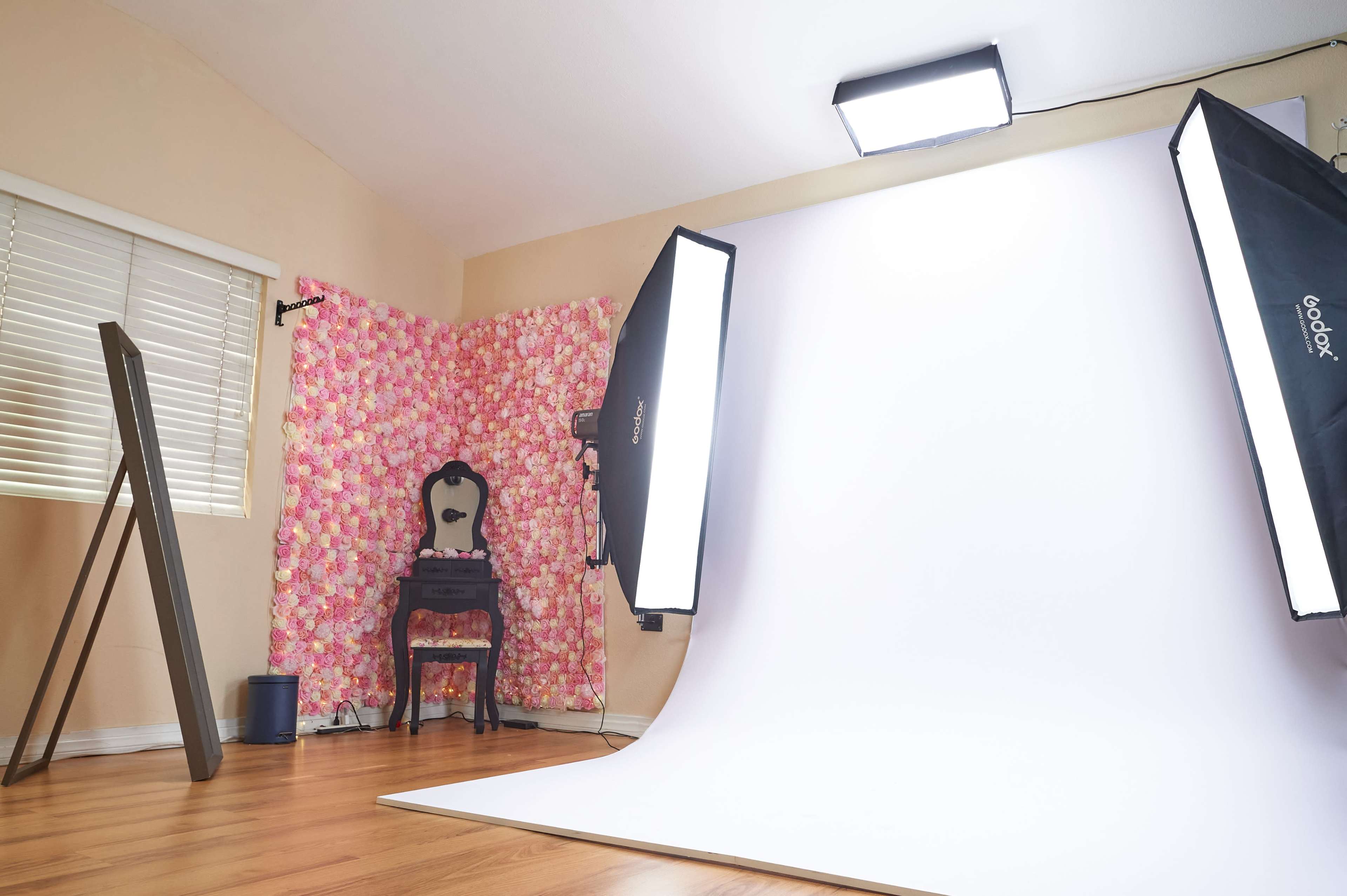 The image depicts a photography studio featuring a backdrop of artificial flowers, a chair, and professional lighting equipment set up for a photoshoot.