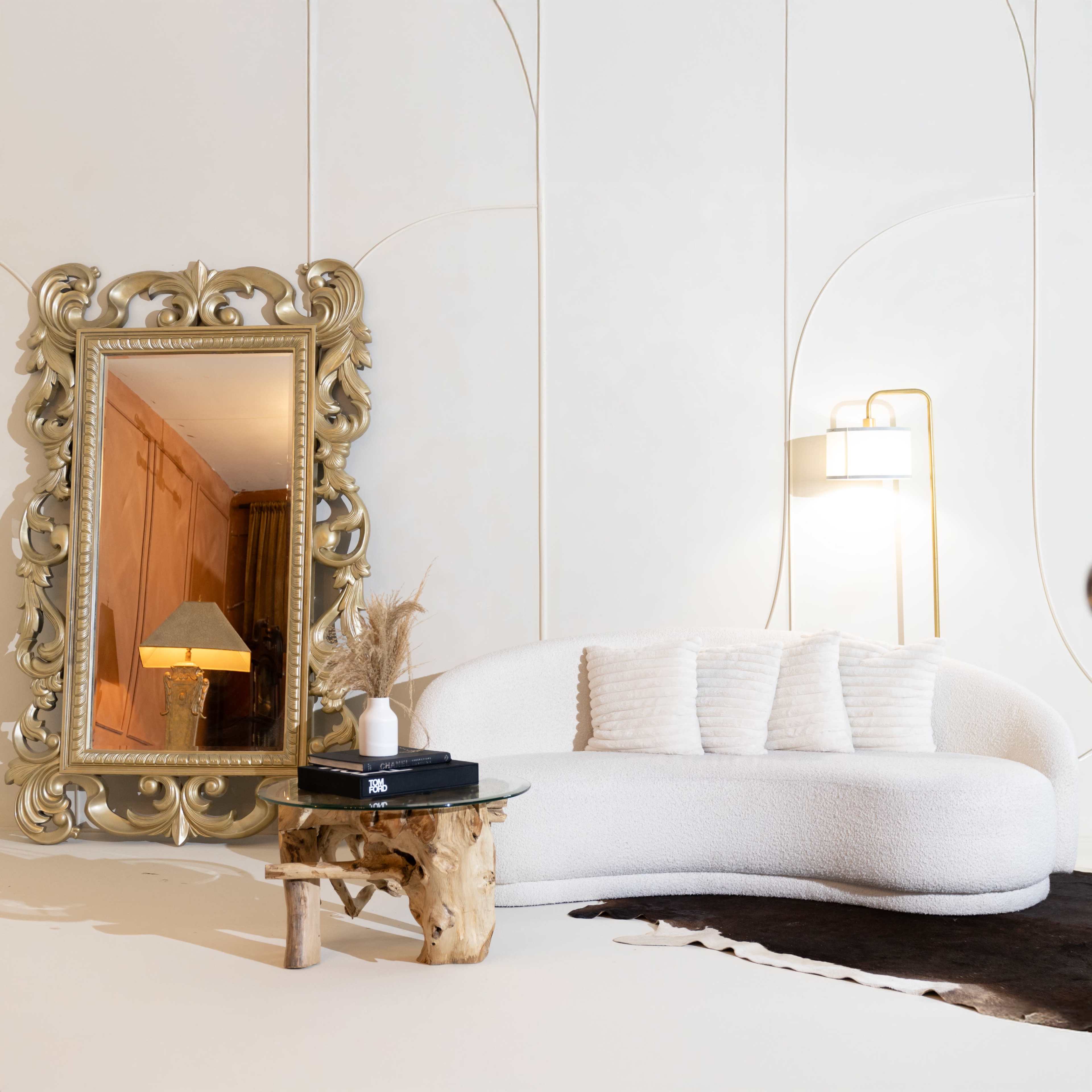 A large ornate mirror stands beside a curved white couch and a wooden table with decorative items in a brightly lit room.