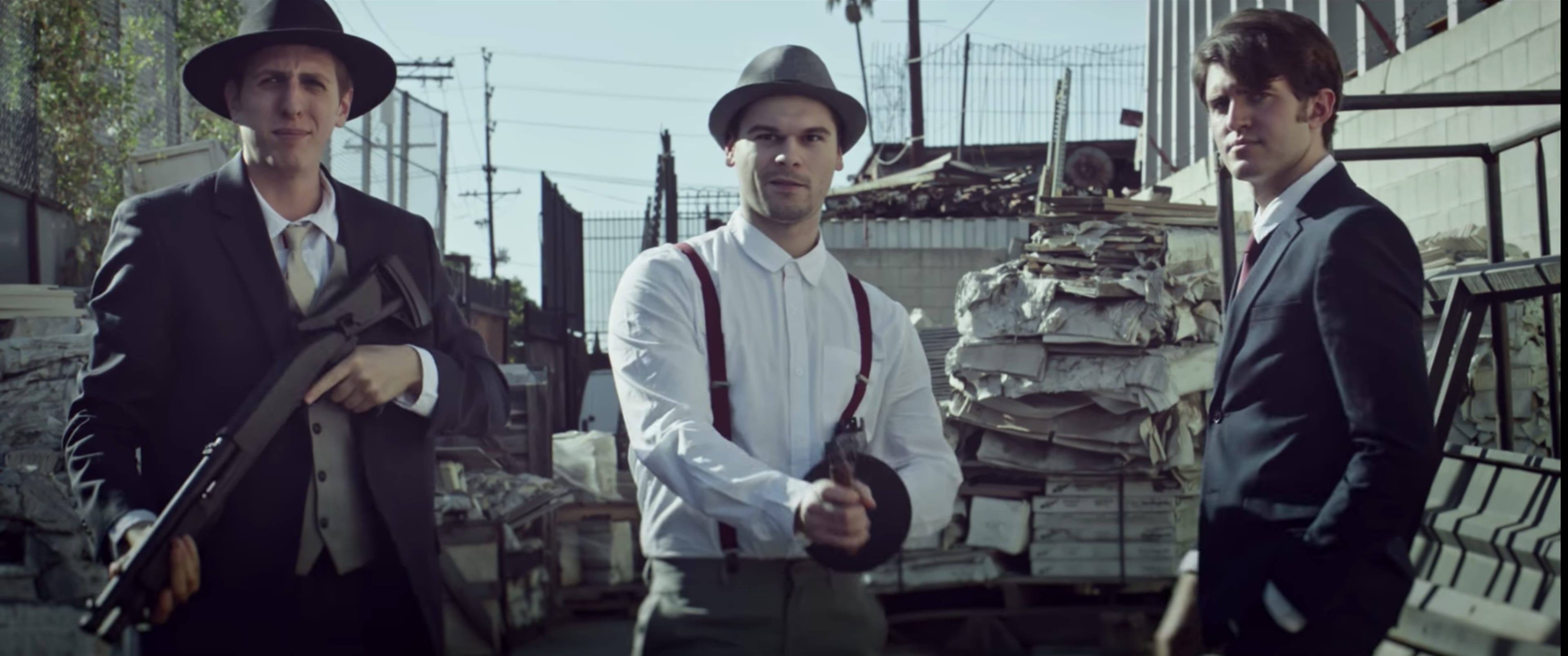 Three men stand in an alleyway surrounded by stacks of papers, with one man holding a gun and the others holding large weapons.