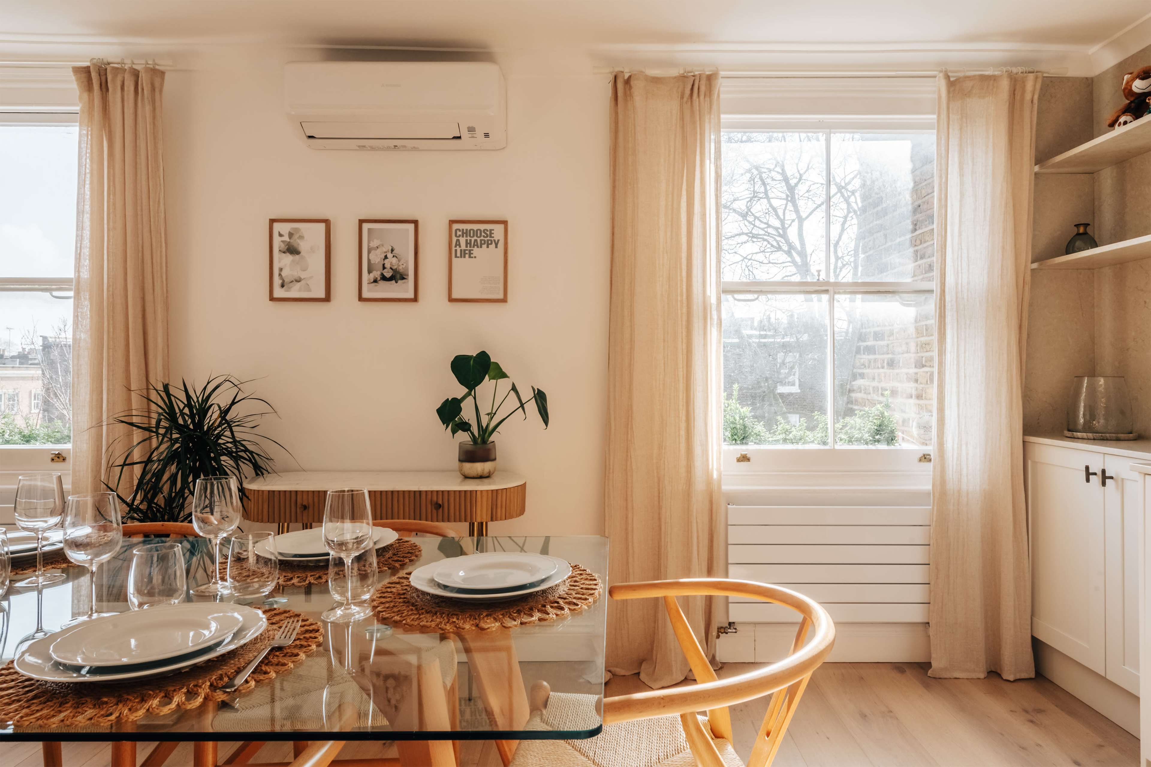 A dining area features a glass table set with dinnerware and glasses, surrounded by wooden chairs, with a plant near the window and framed pictures on the wall.