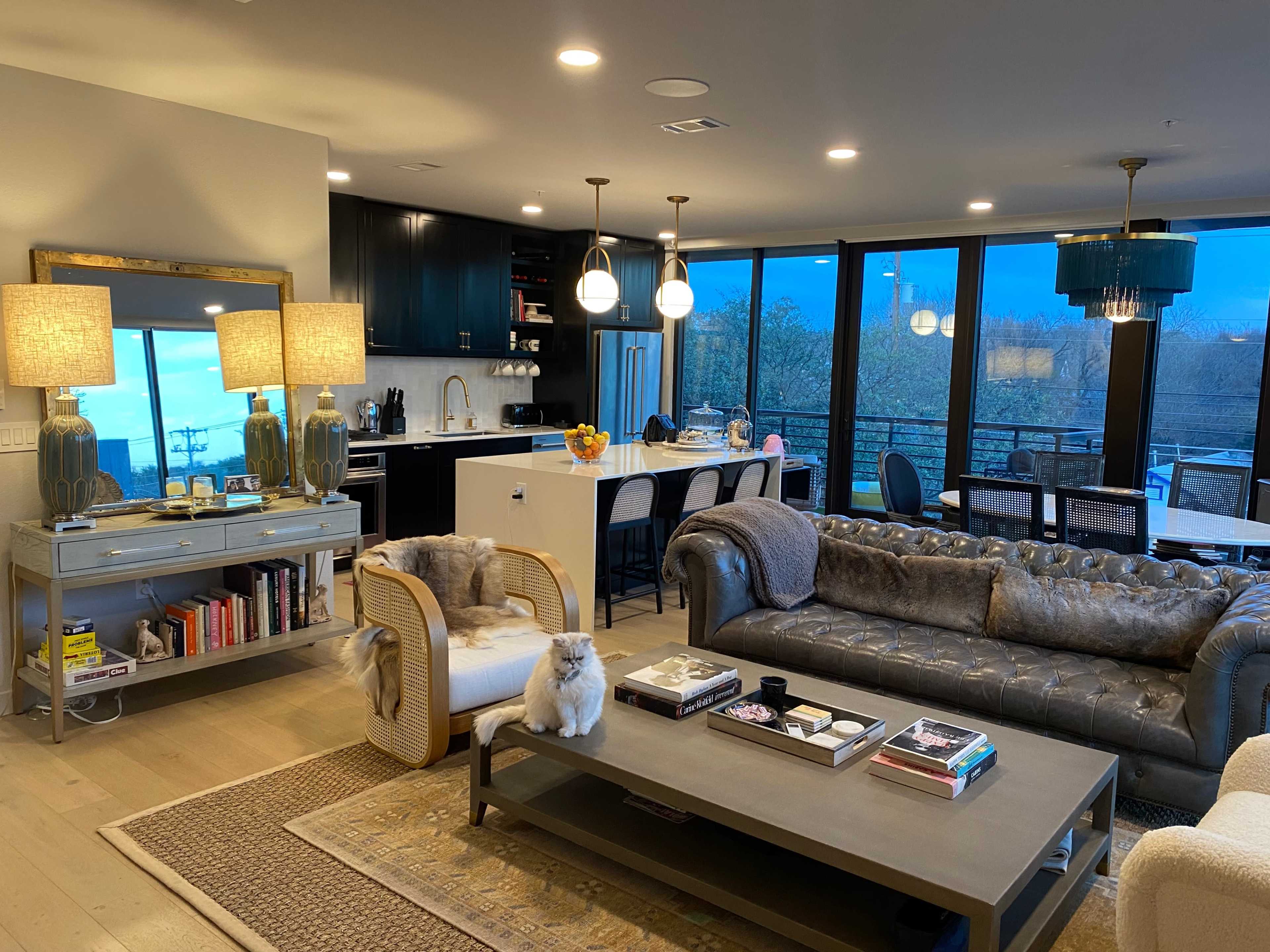 The image shows a modern living room with a sofa, coffee table, and decorative lamps, alongside a view of an outdoor area through large glass windows.