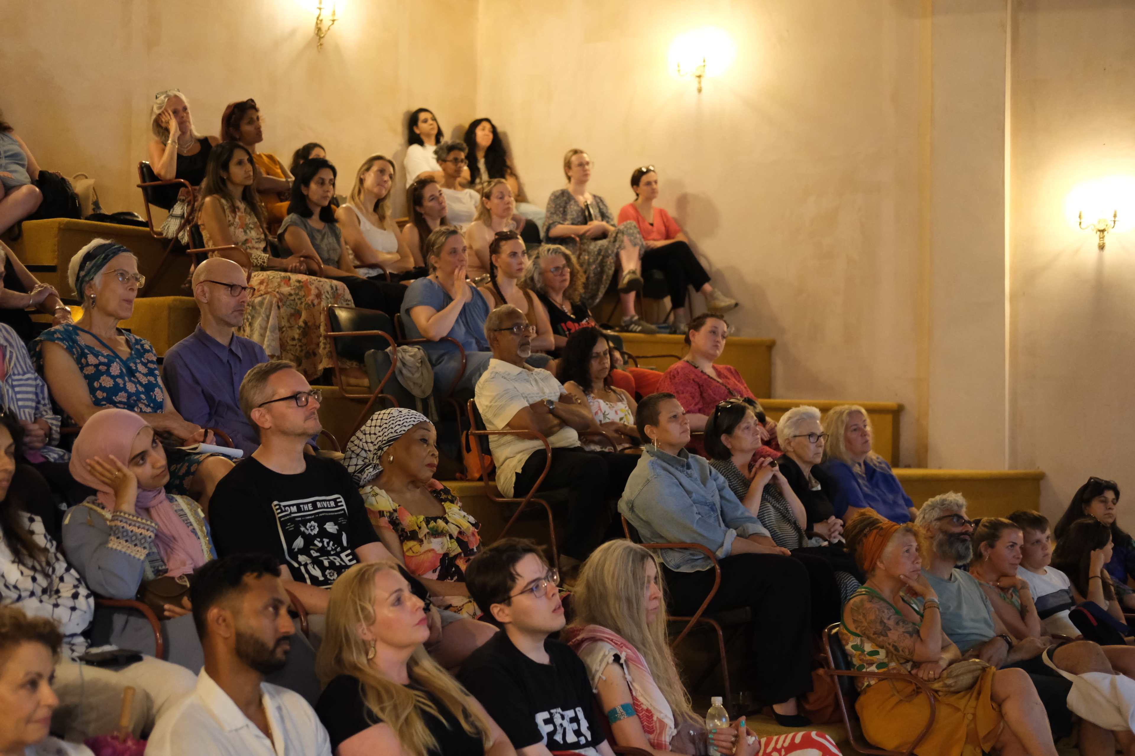 A diverse audience watches a presentation in a tiered seating arrangement within a softly lit room.