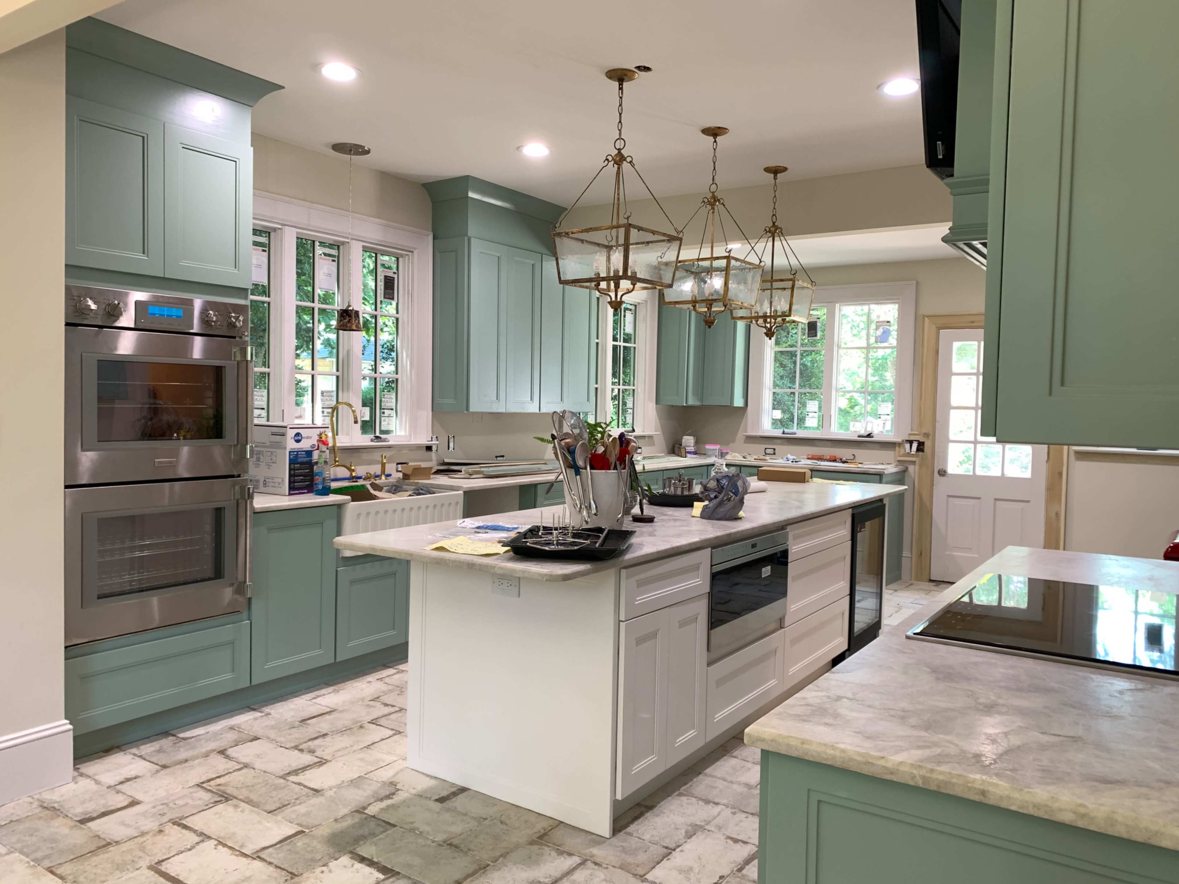 A modern kitchen featuring light green cabinetry, a central island with a countertop, and multiple light fixtures hanging above.
