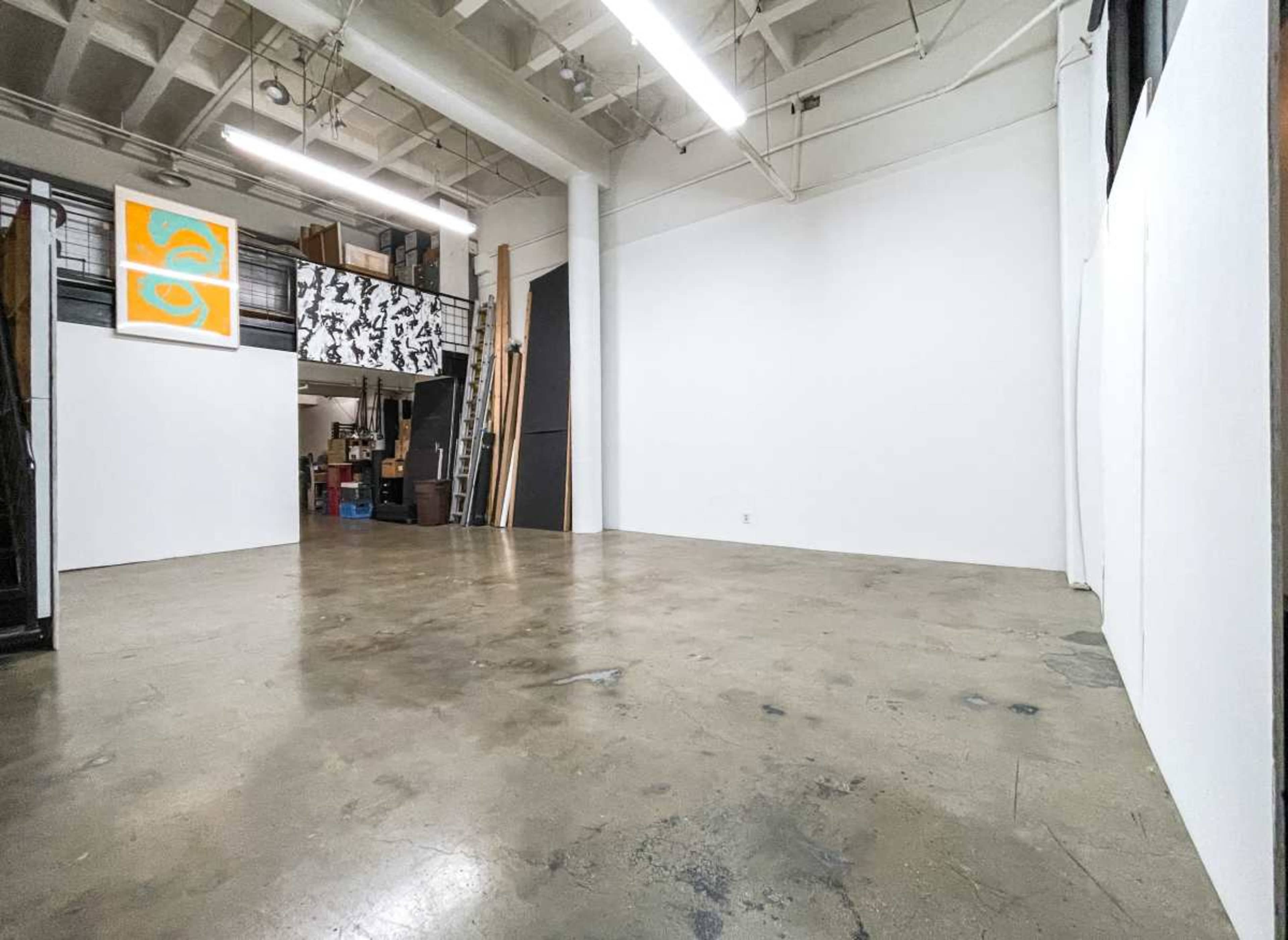 The image shows an empty gallery space with a polished concrete floor, white walls, and a staircase in the background.