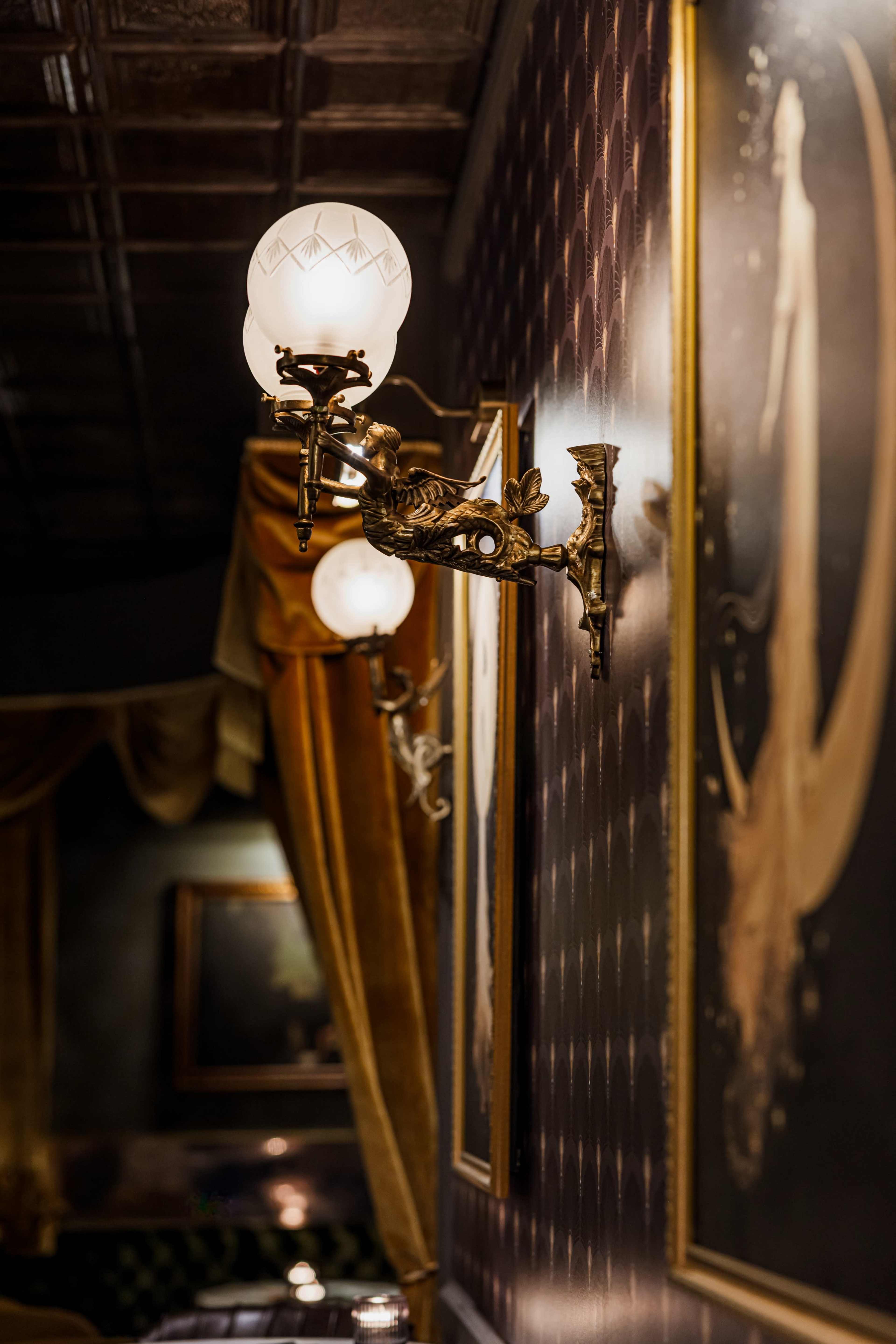 The image shows elegant wall sconces with glass globes, illuminating a tastefully decorated interior featuring dark patterned walls and framed artworks.