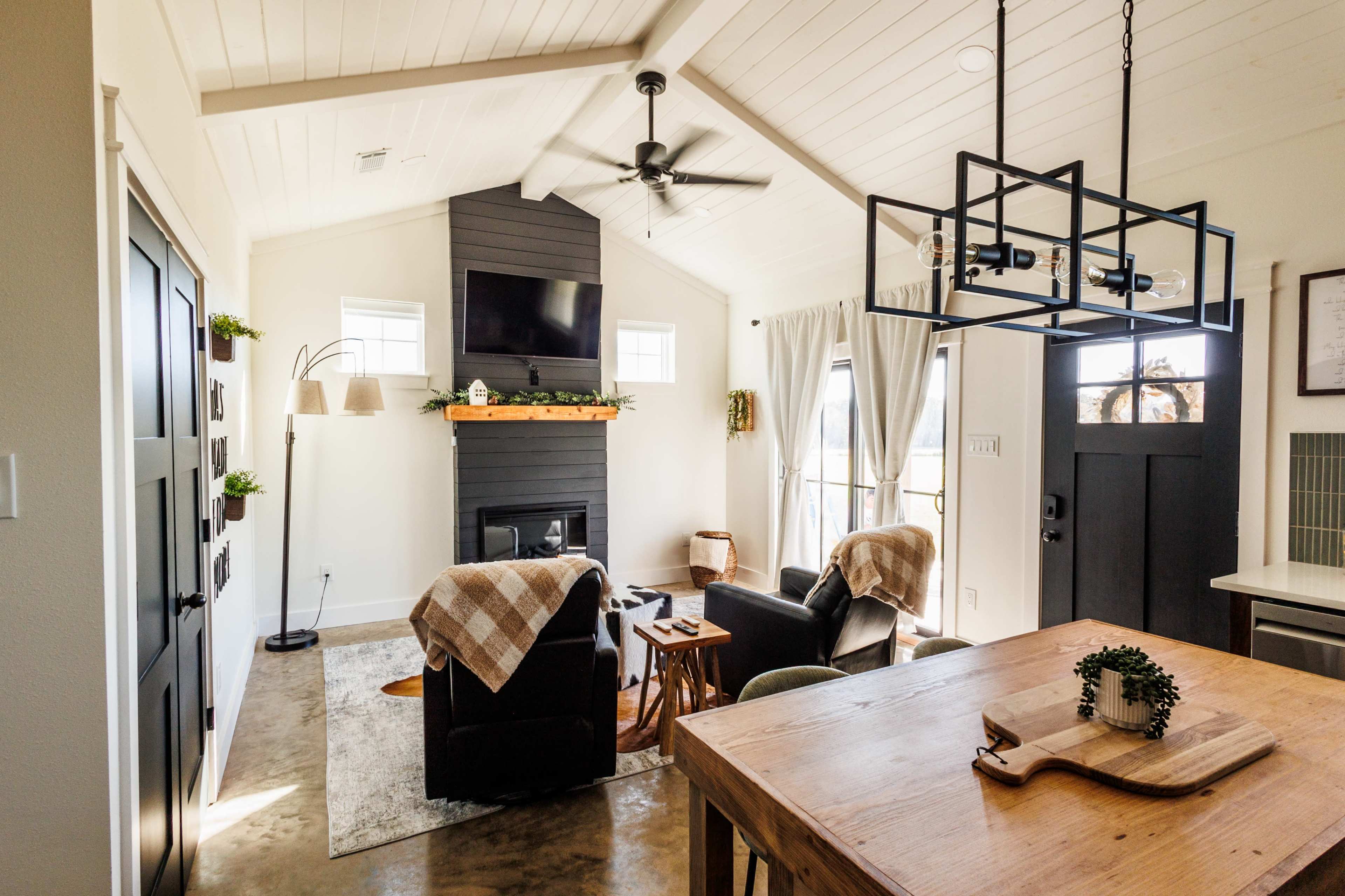 The image shows a modern living room with a vaulted ceiling, a black fireplace, and a dining area featuring a wooden table and a lantern-style chandelier.