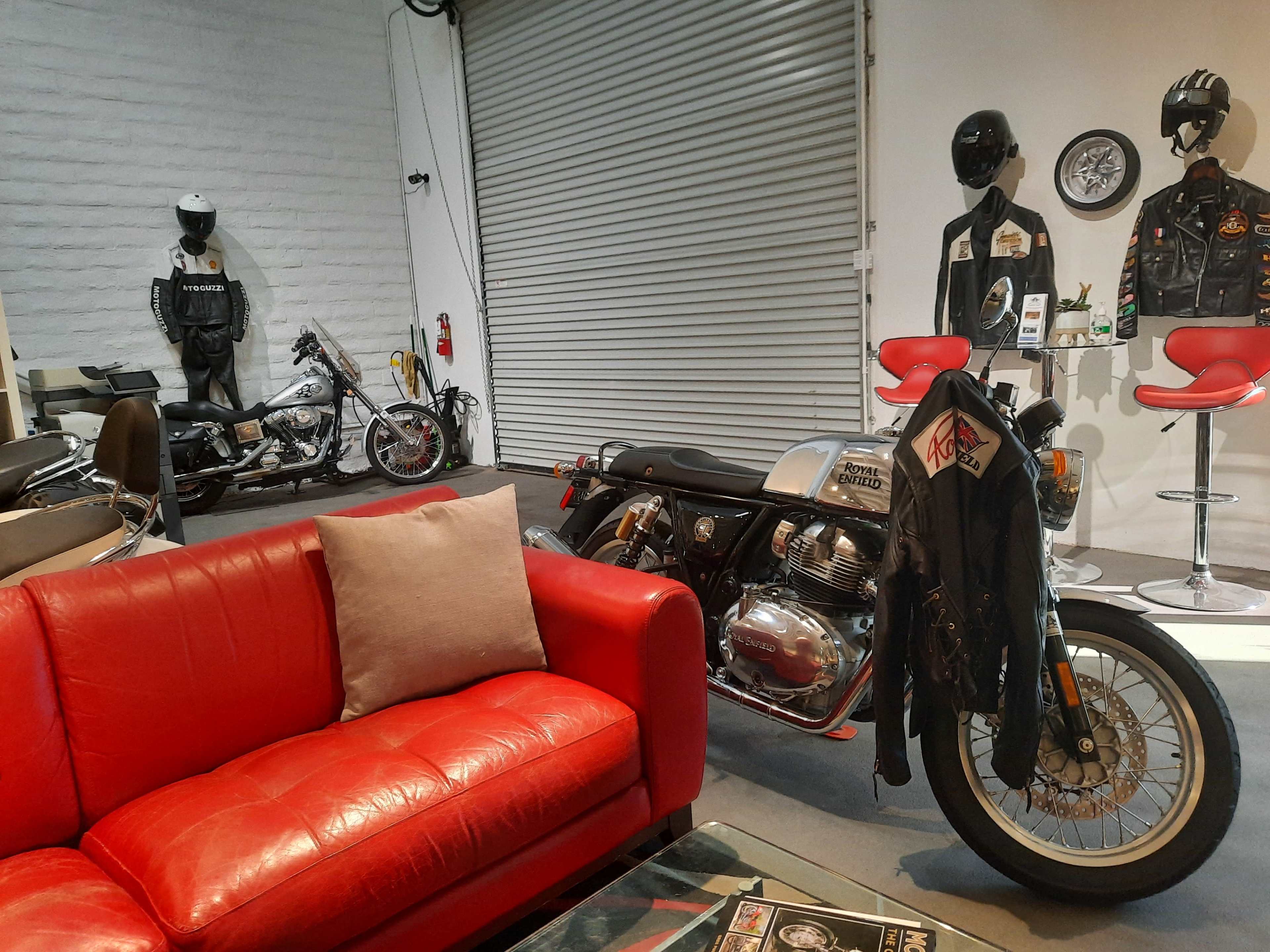 The scene shows a modern garage or showroom with a red couch, two motorcycles, mannequins dressed in motorcycle gear, and a bar-style seating area.