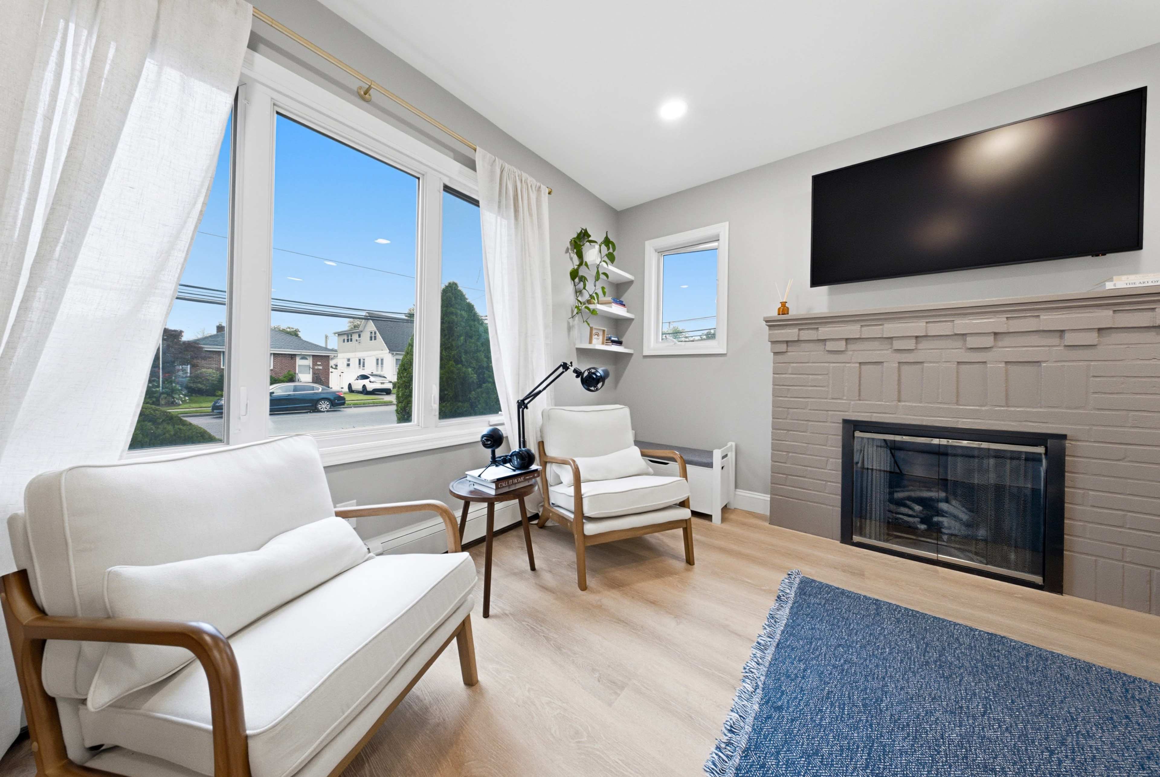A cozy living room features a fireplace, two white armchairs, a mounted television, and large windows letting in natural light.