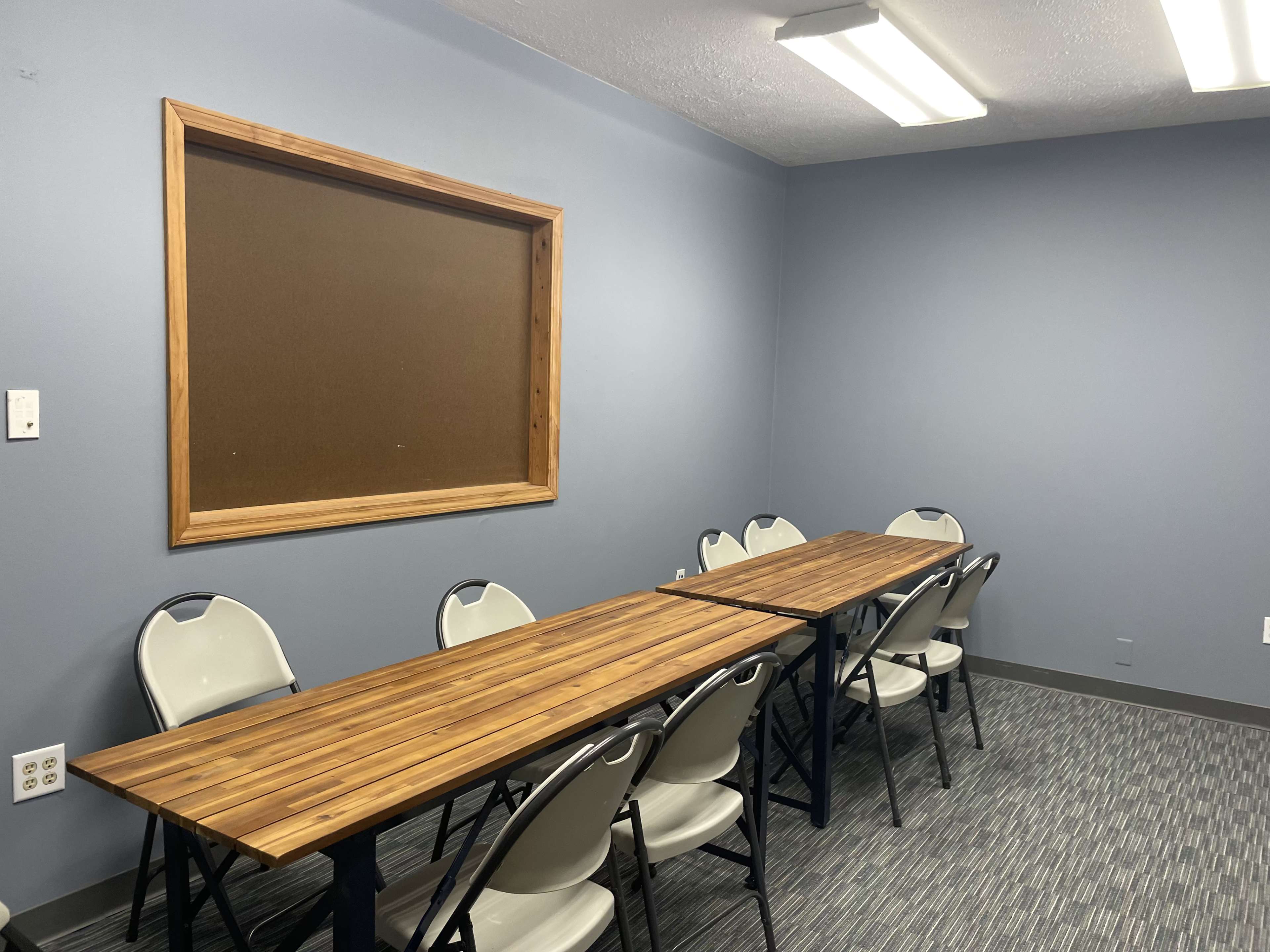 A meeting room with two long wooden tables facing each other, surrounded by folding chairs, and a large framed bulletin board on the wall.