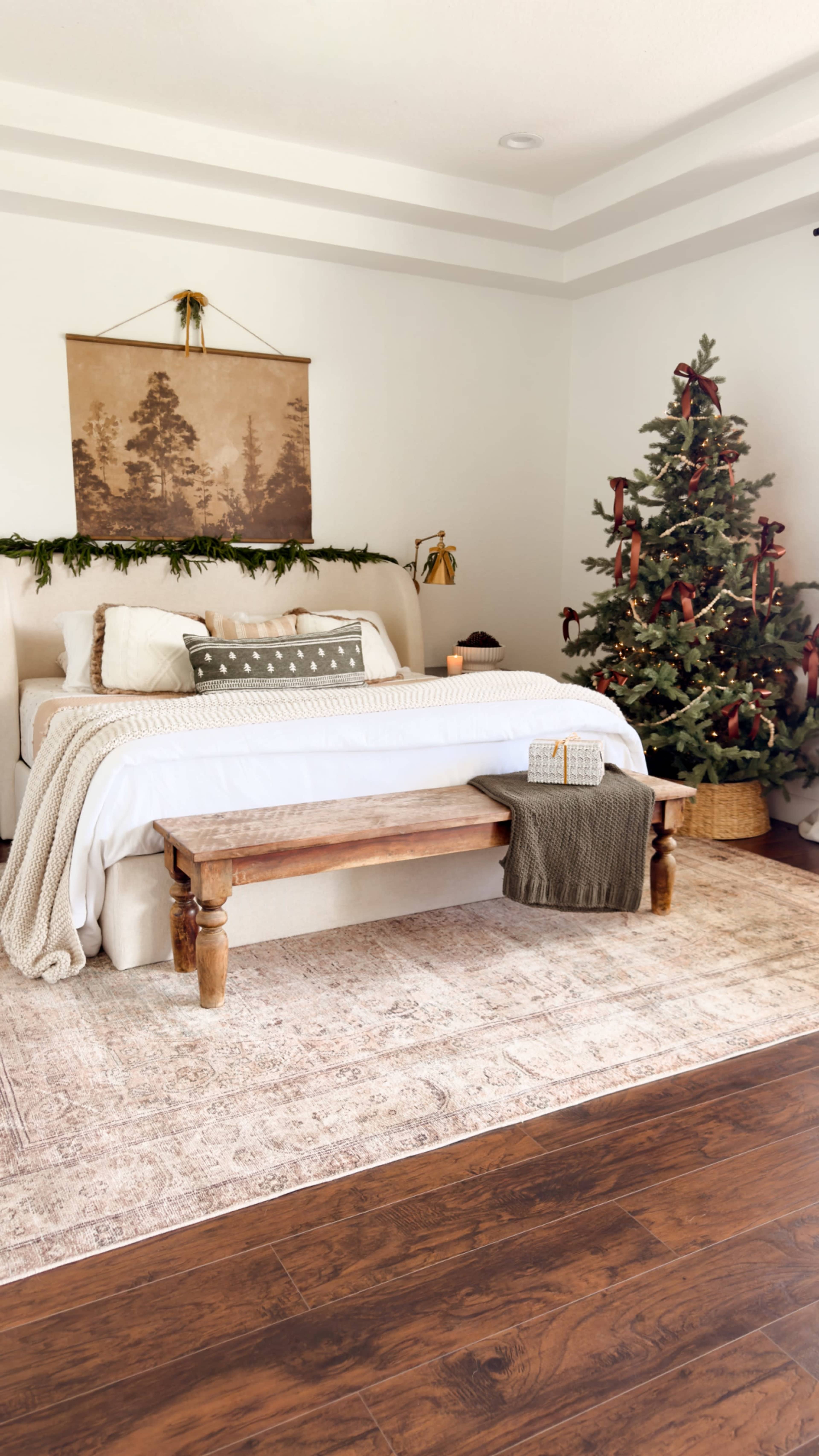 The image shows a cozy bedroom with a bed adorned with a white comforter and decorative pillows, a wooden bench at the foot, and a Christmas tree decorated in the corner.