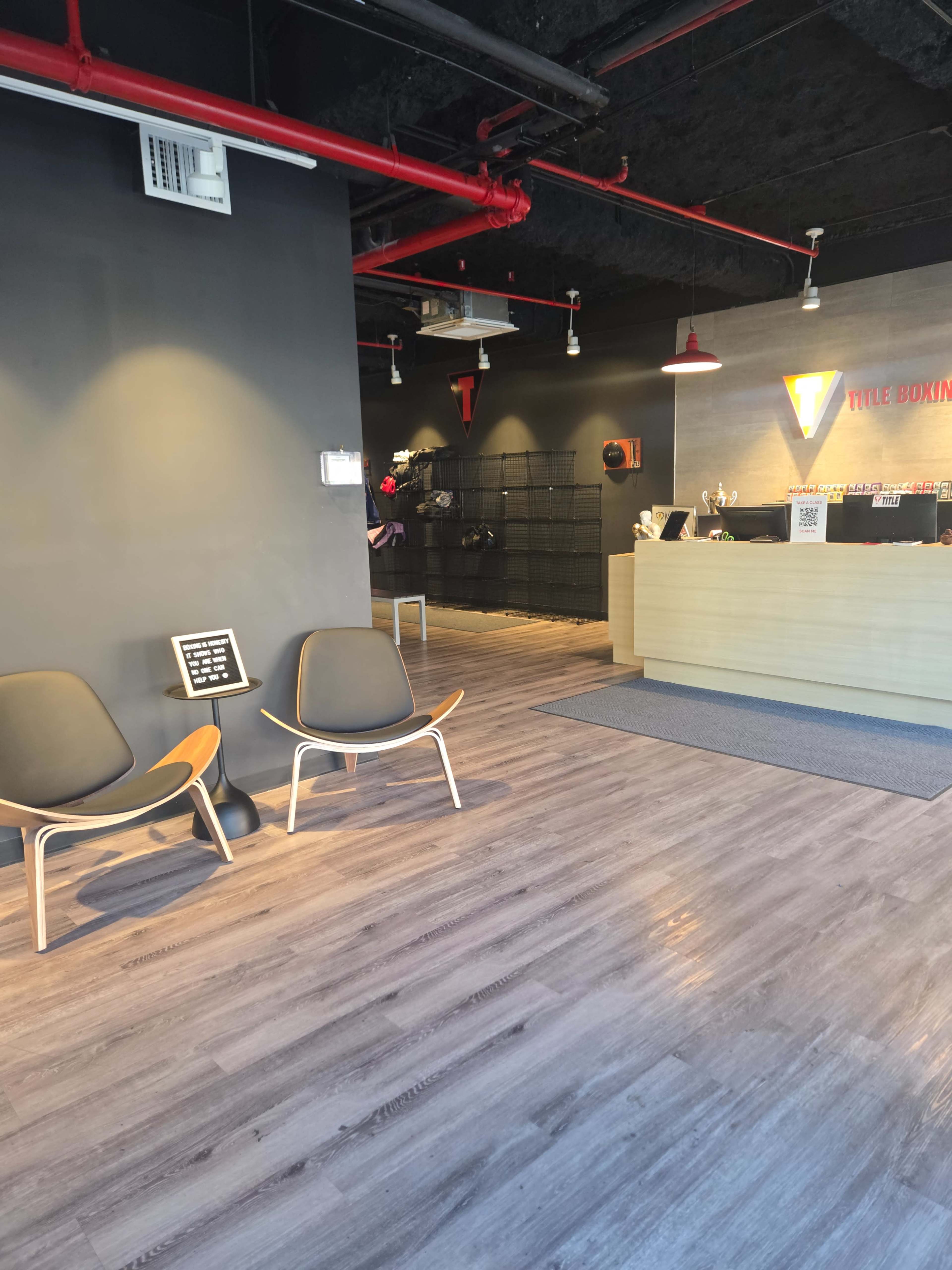 The image shows a modern reception area of a fitness studio with wooden flooring, two black chairs, and a front desk.