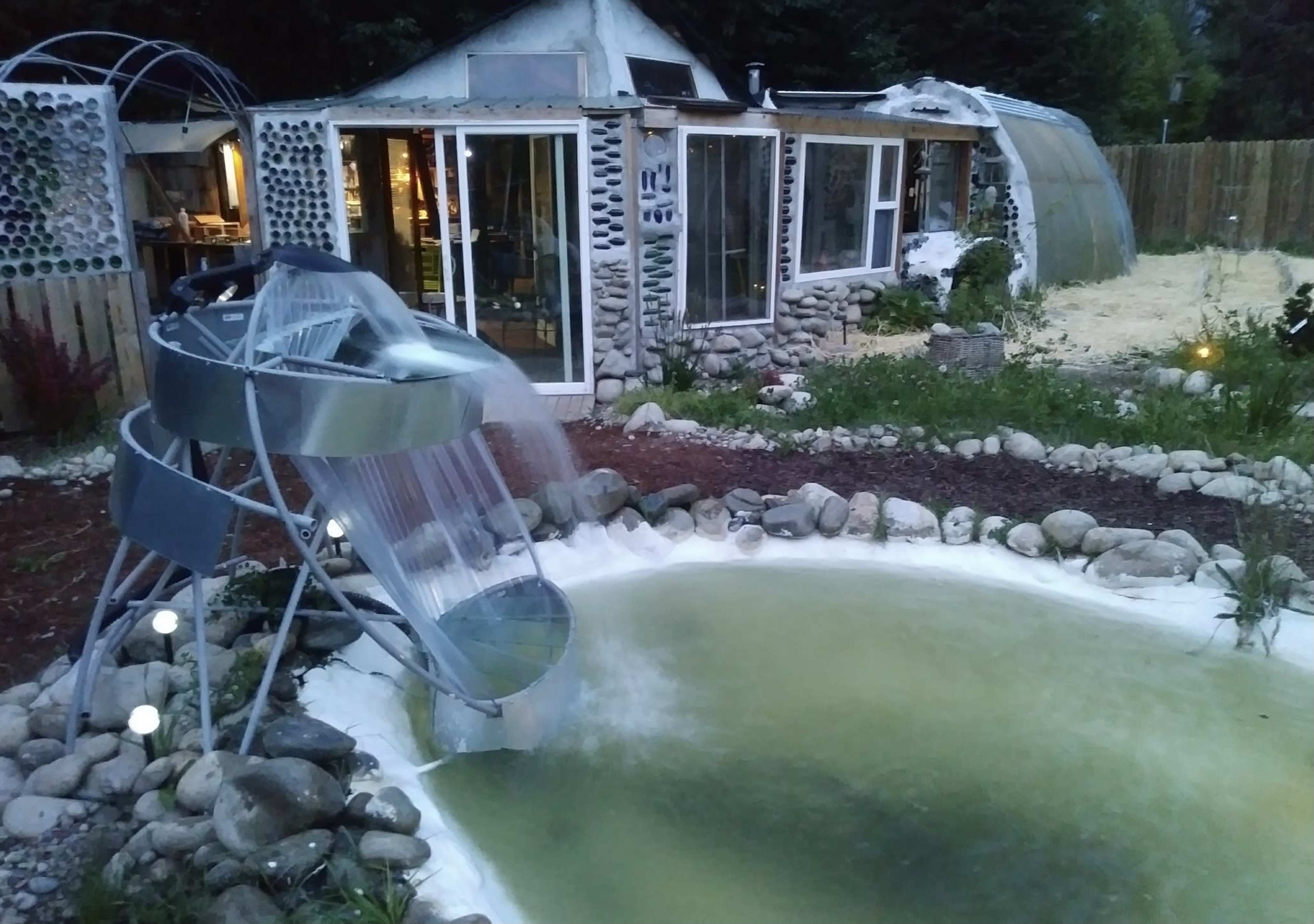 A modern water slide feeding into a small pond, adjacent to a house made of stone and glass, surrounded by a garden.
