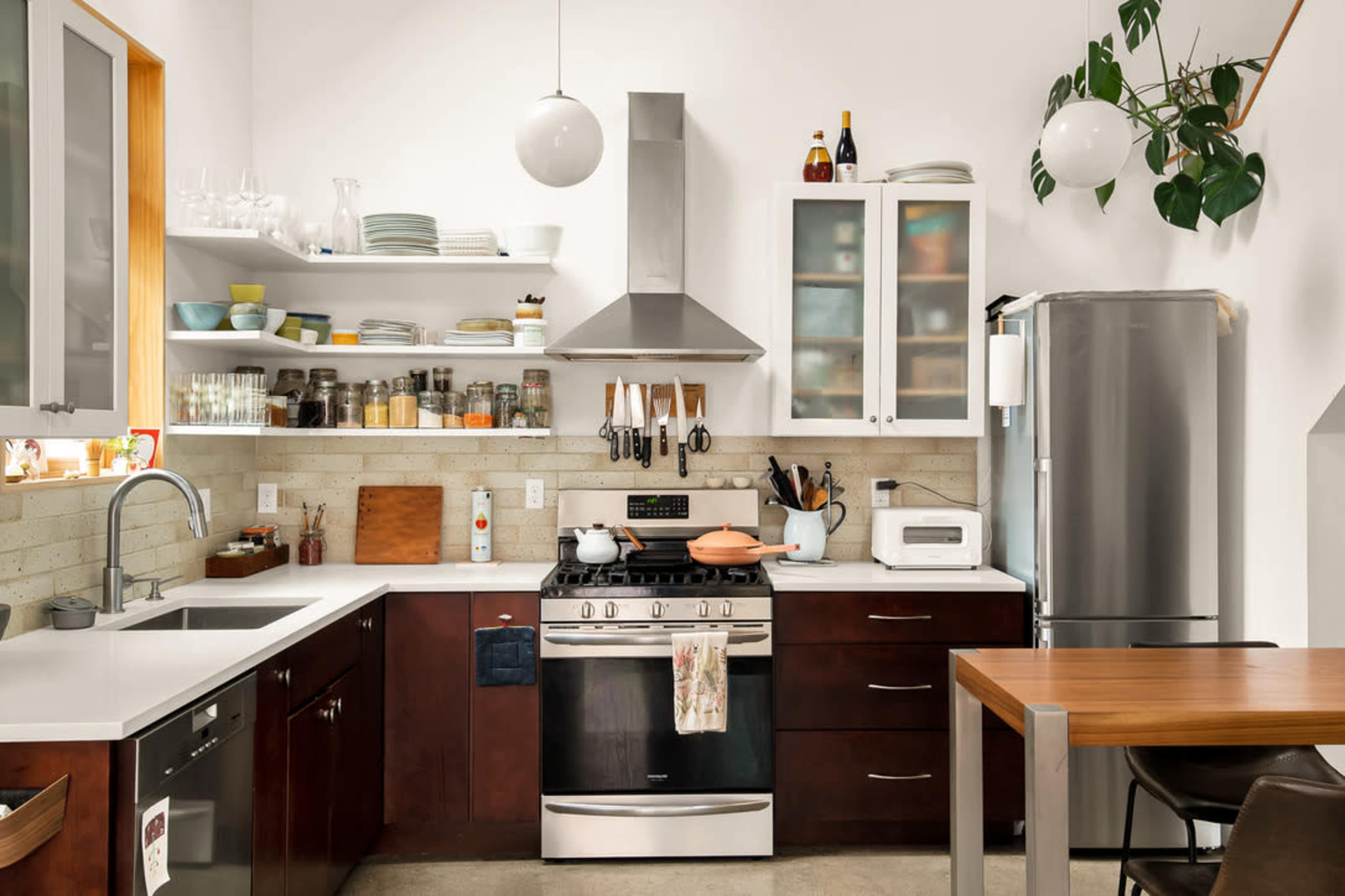 A modern kitchen features stainless steel appliances, wooden cabinets, open shelving with dishware, and a dining table.