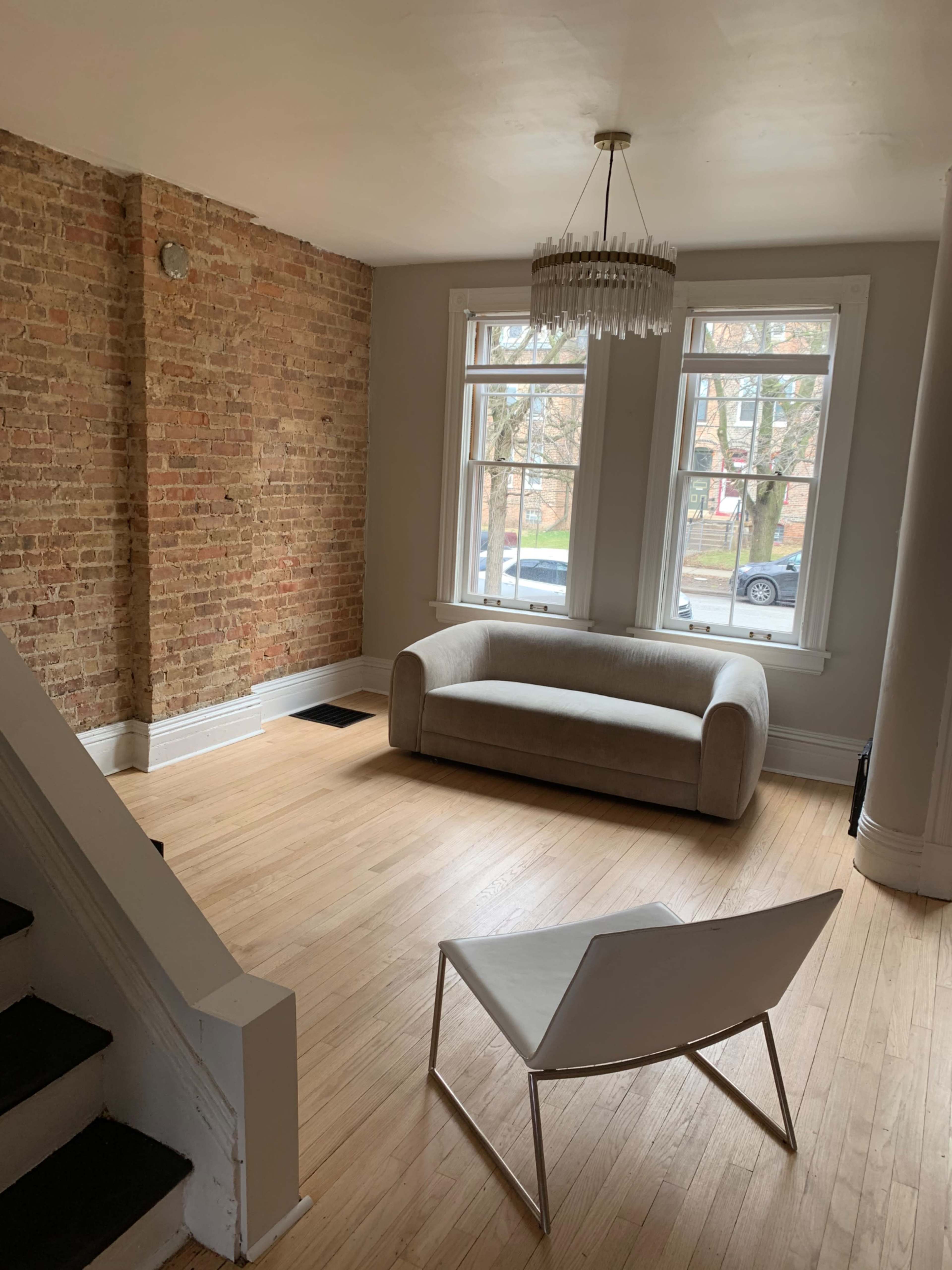 A sparsely furnished living room featuring a brick wall, a light-colored sofa, a white chair, and large windows that overlook a street.