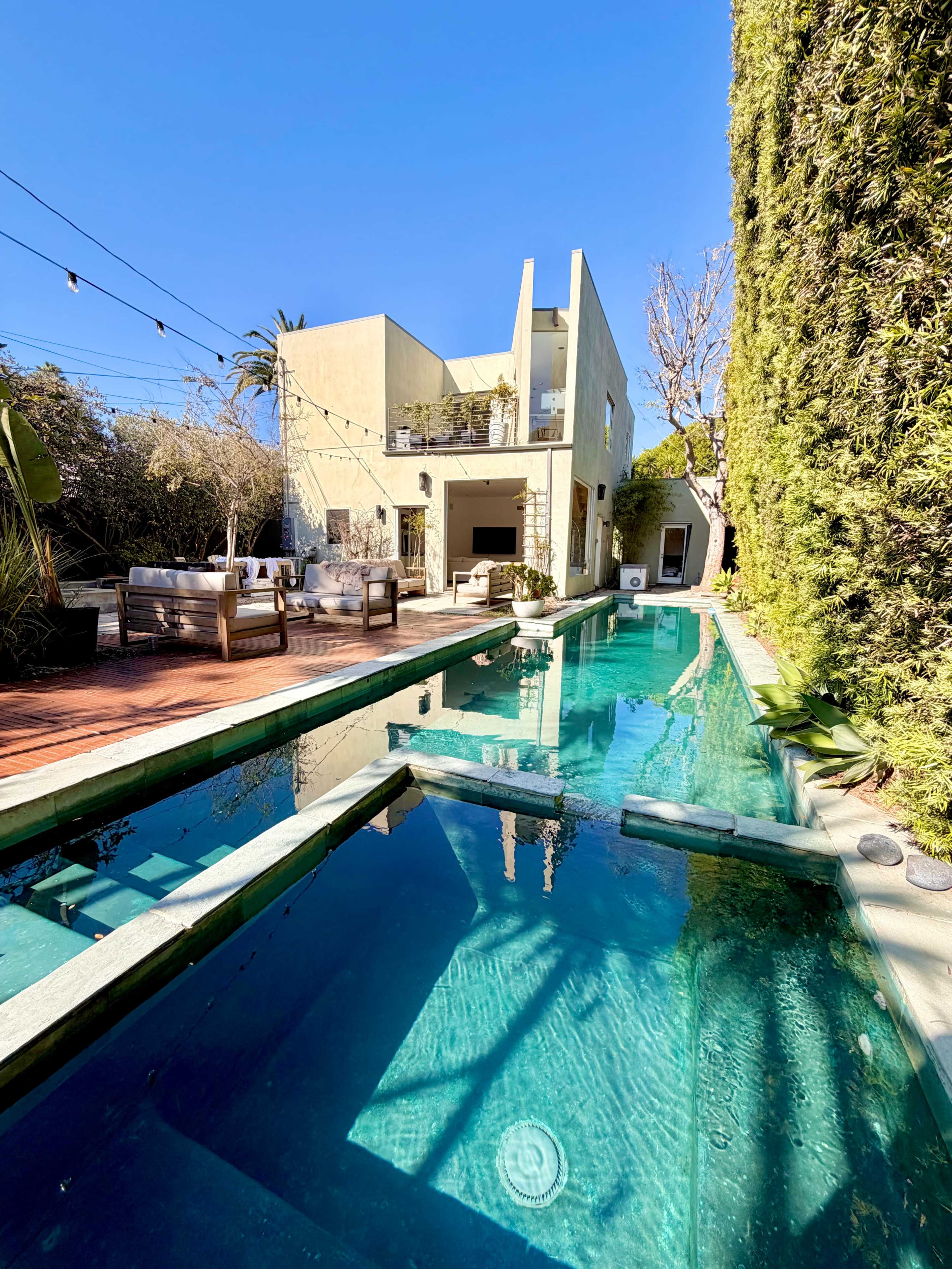 The image shows a modern house with a swimming pool surrounded by outdoor seating and tall greenery under a clear blue sky.