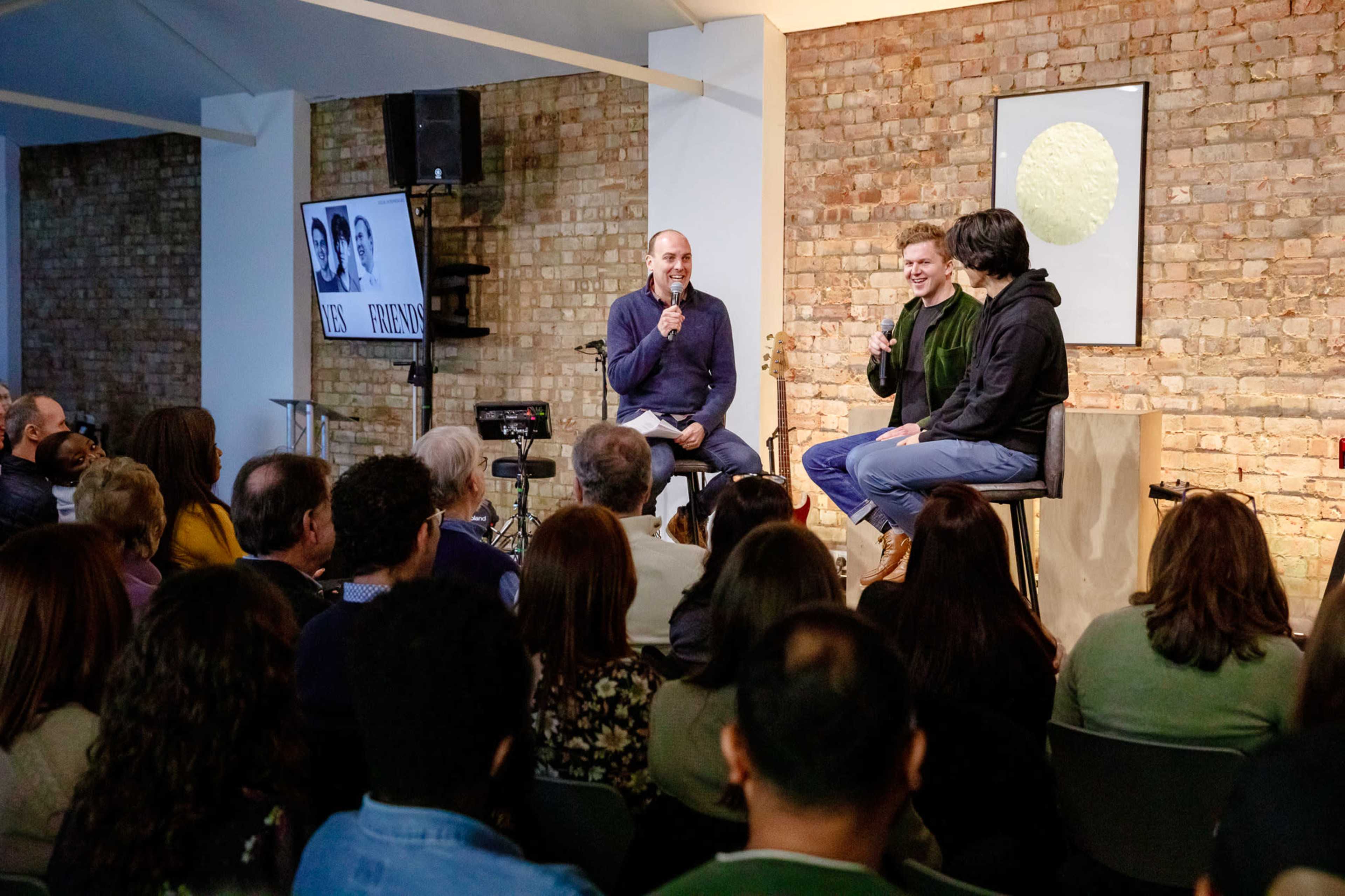 A panel discussion with three speakers takes place in front of an audience seated in a brick-walled venue.