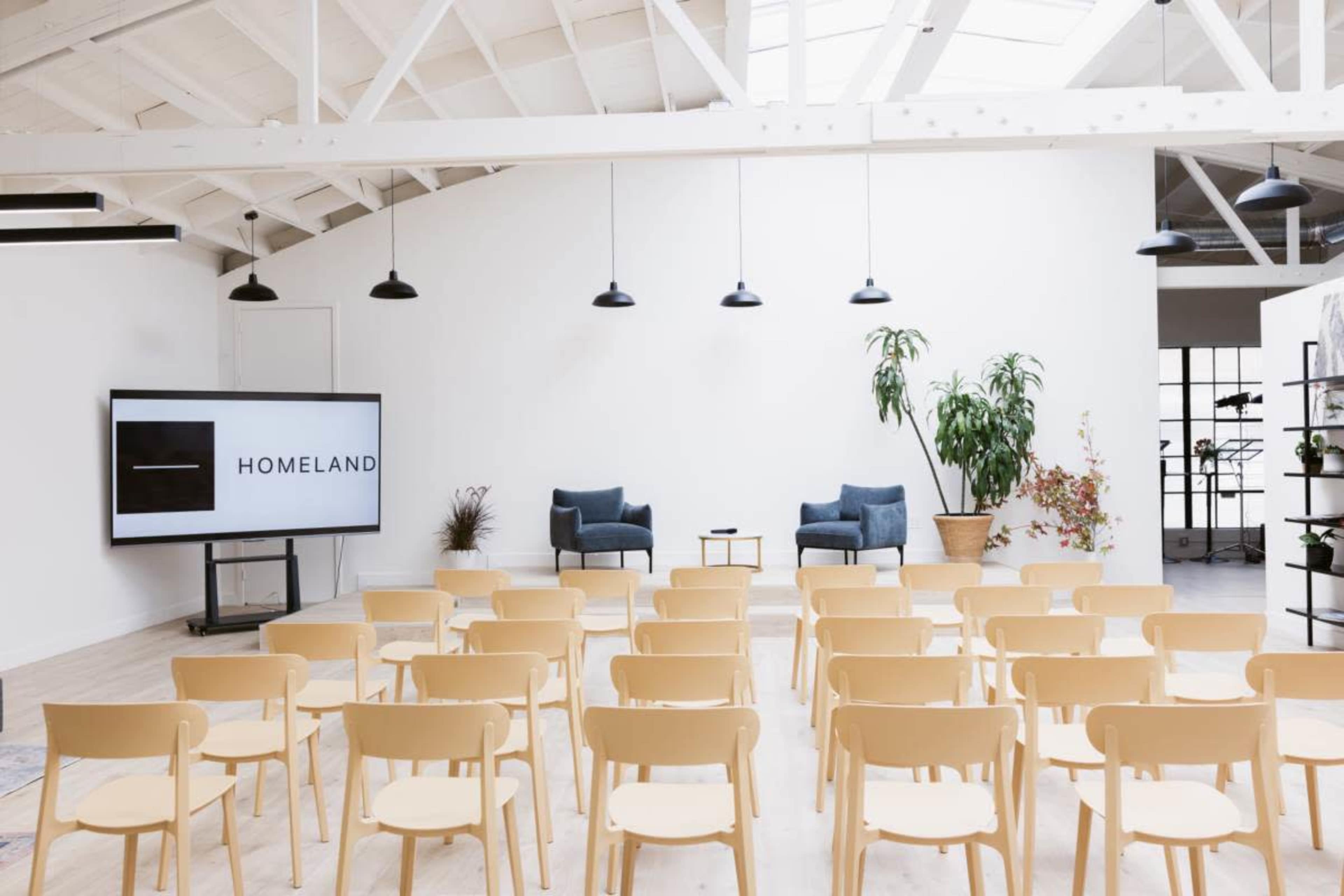The image shows a spacious room arranged for an event, featuring rows of wooden chairs, a large screen displaying the word "HOMELAND," and two blue armchairs beside a small table.