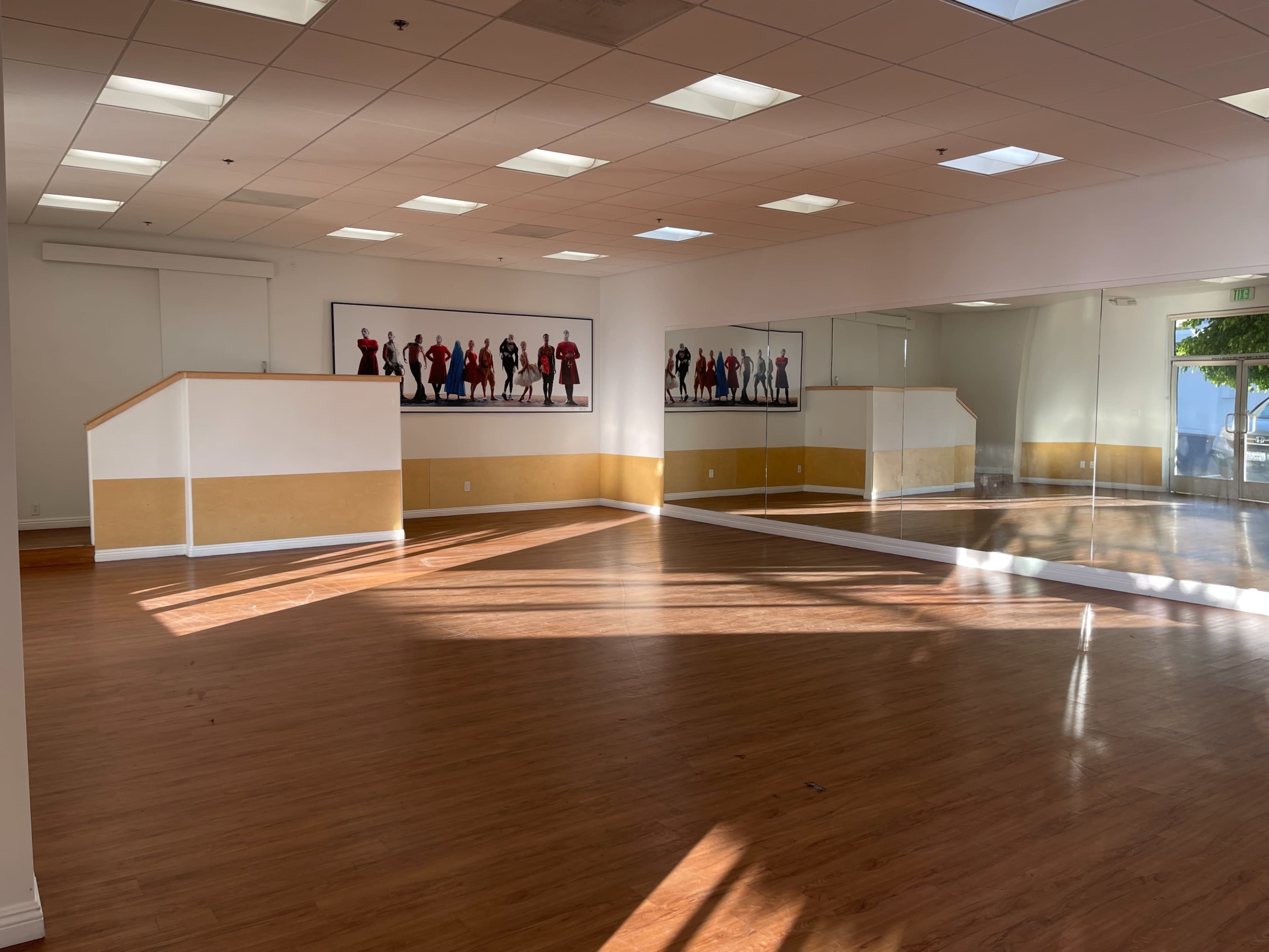 The image shows a spacious and well-lit dance studio with wooden flooring, large windows, and a mirror on one wall.