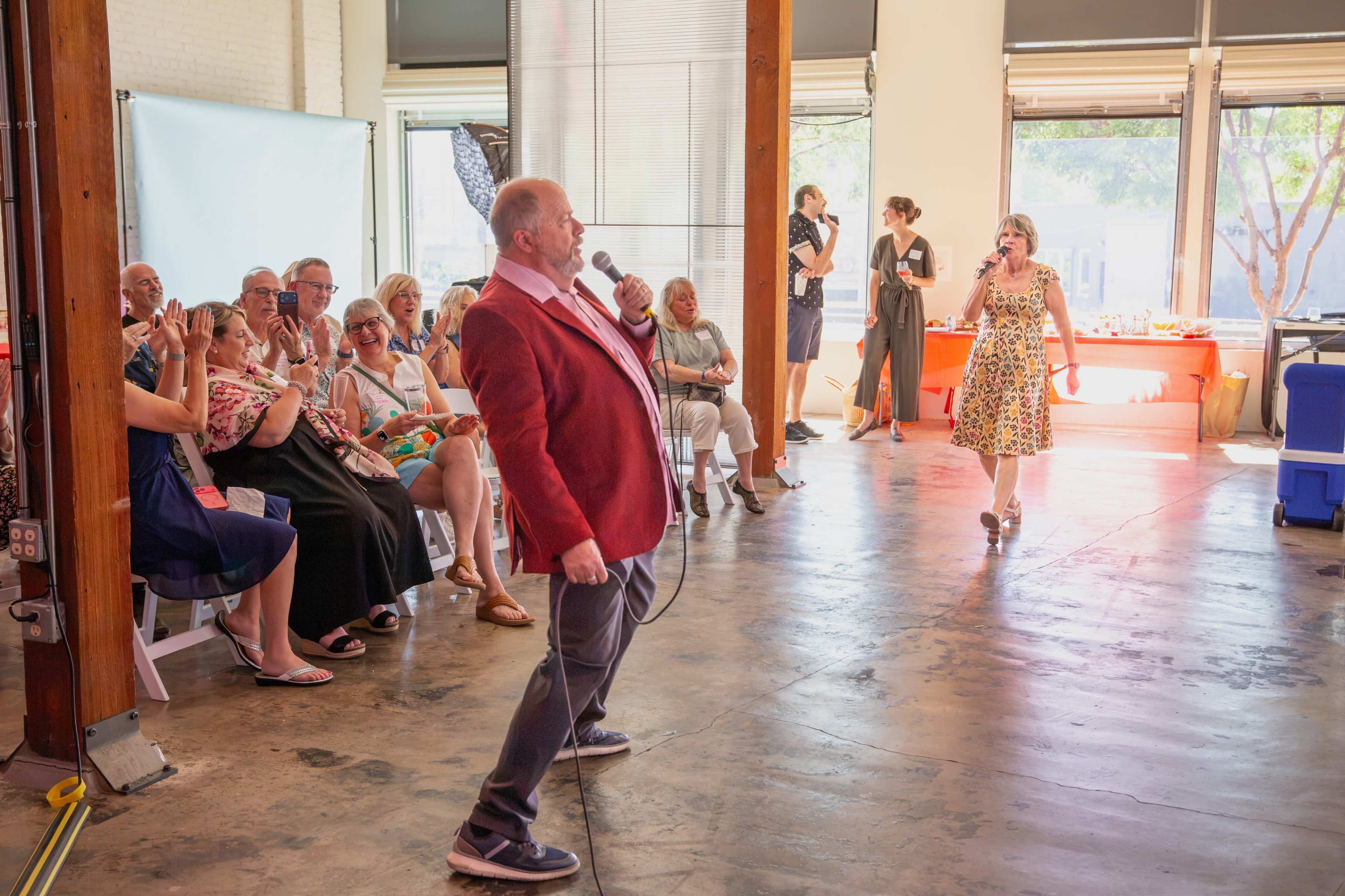A man in a red blazer performs with a microphone while an audience of seated guests watches in a spacious indoor venue.