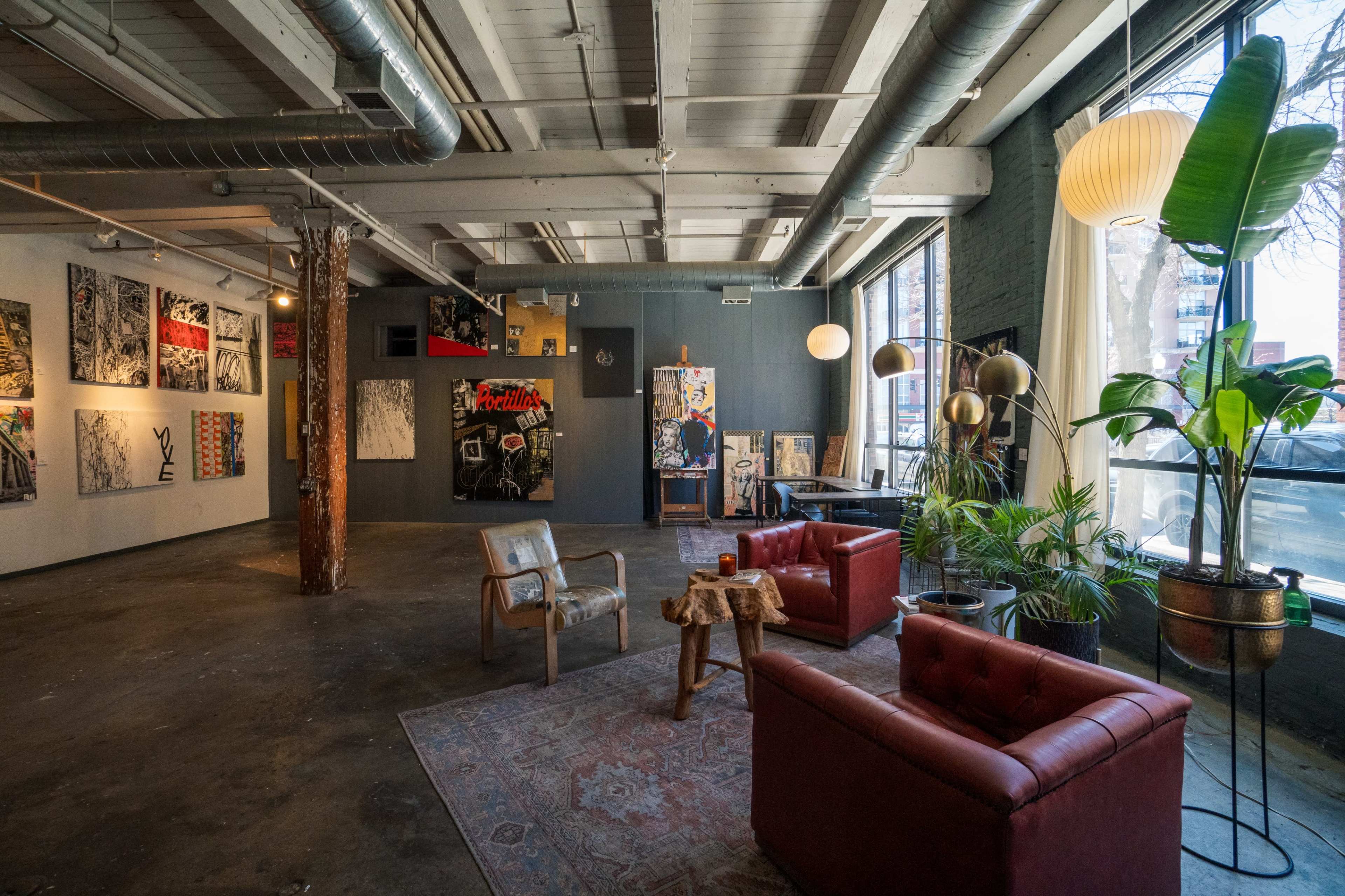 A spacious art gallery with exposed beams, featuring various artwork on the walls, red leather chairs, and decorative plants.