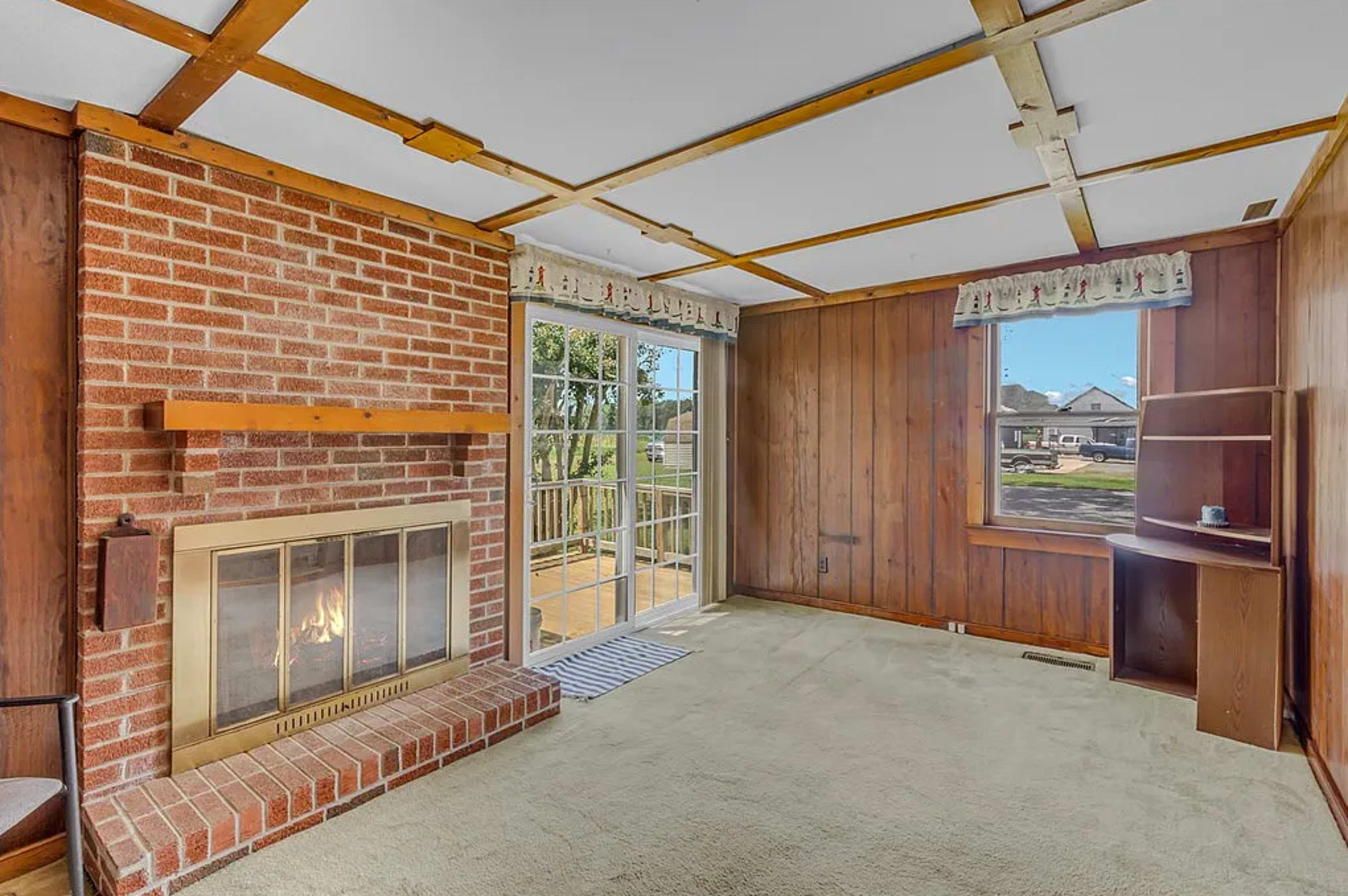 The image shows a room with a brick fireplace, a large window overlooking a yard, and wooden paneling on the walls.