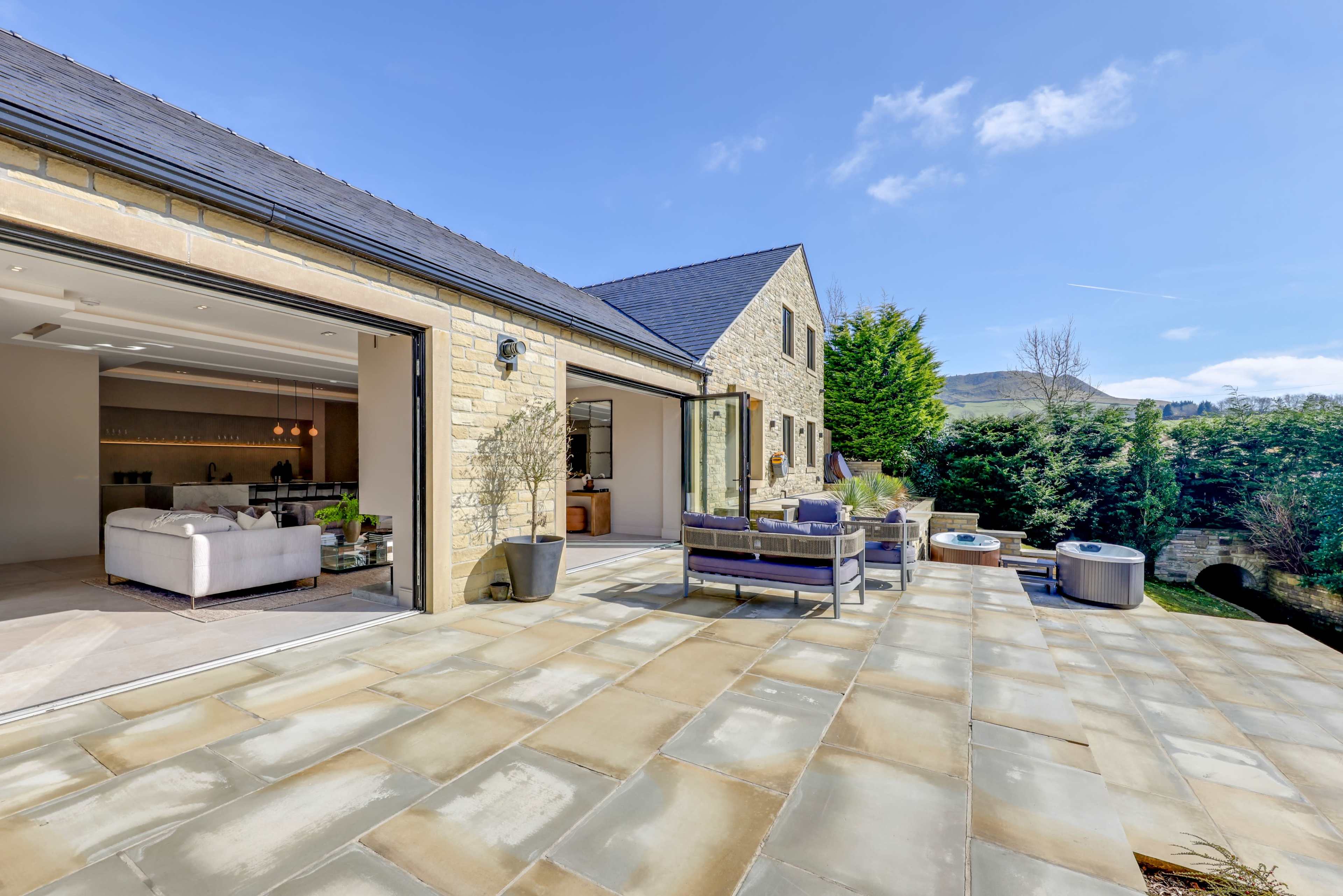 The image shows a stone house with an open patio area featuring seating and views of greenery and hills in the background.