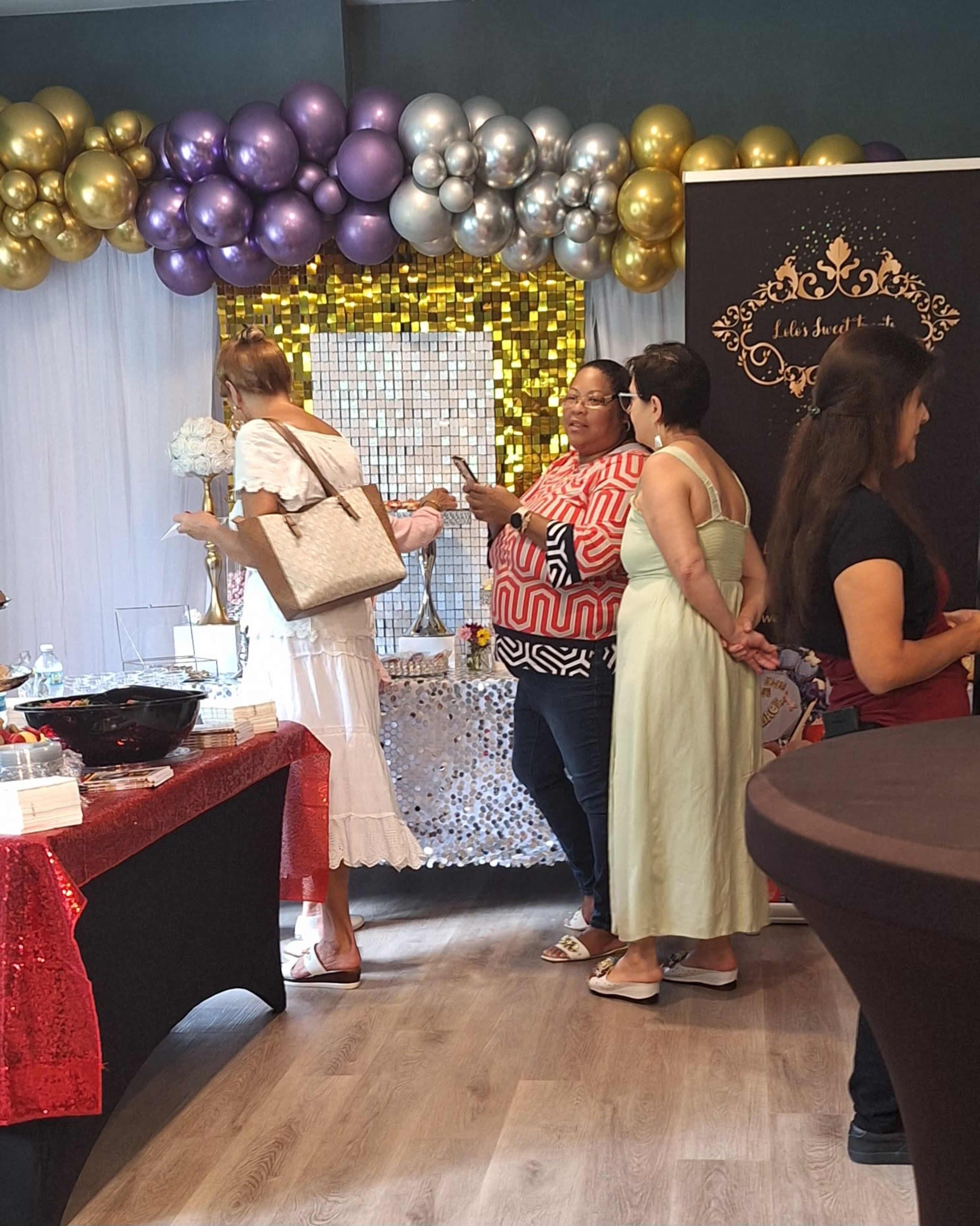 A gathering with attendees socializing in front of a backdrop of balloons and decorations, with tables covered in colorful cloth.