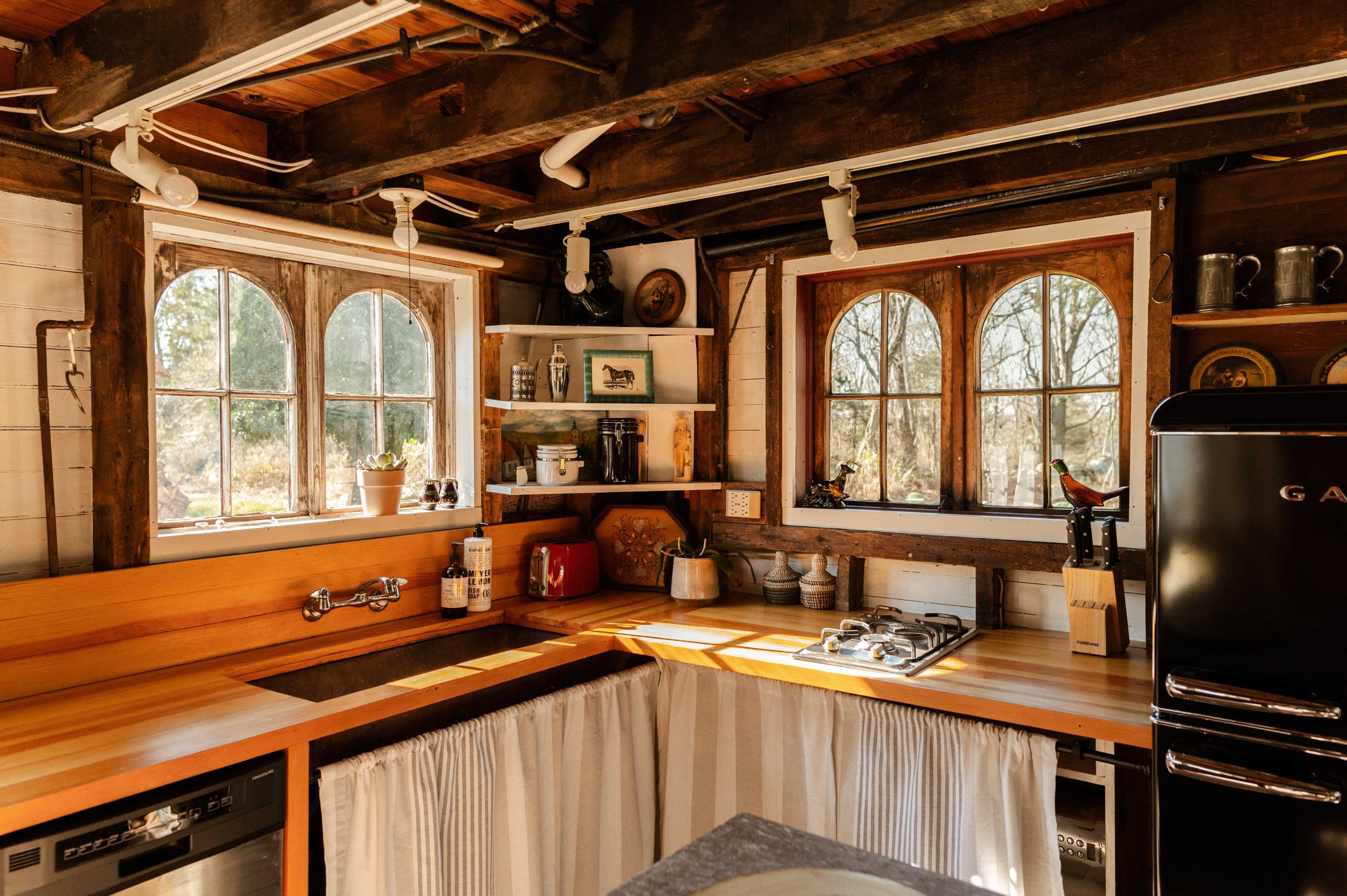 The image shows a rustic kitchen featuring wooden countertops, a gas stove, and two arched windows with natural light streaming in.