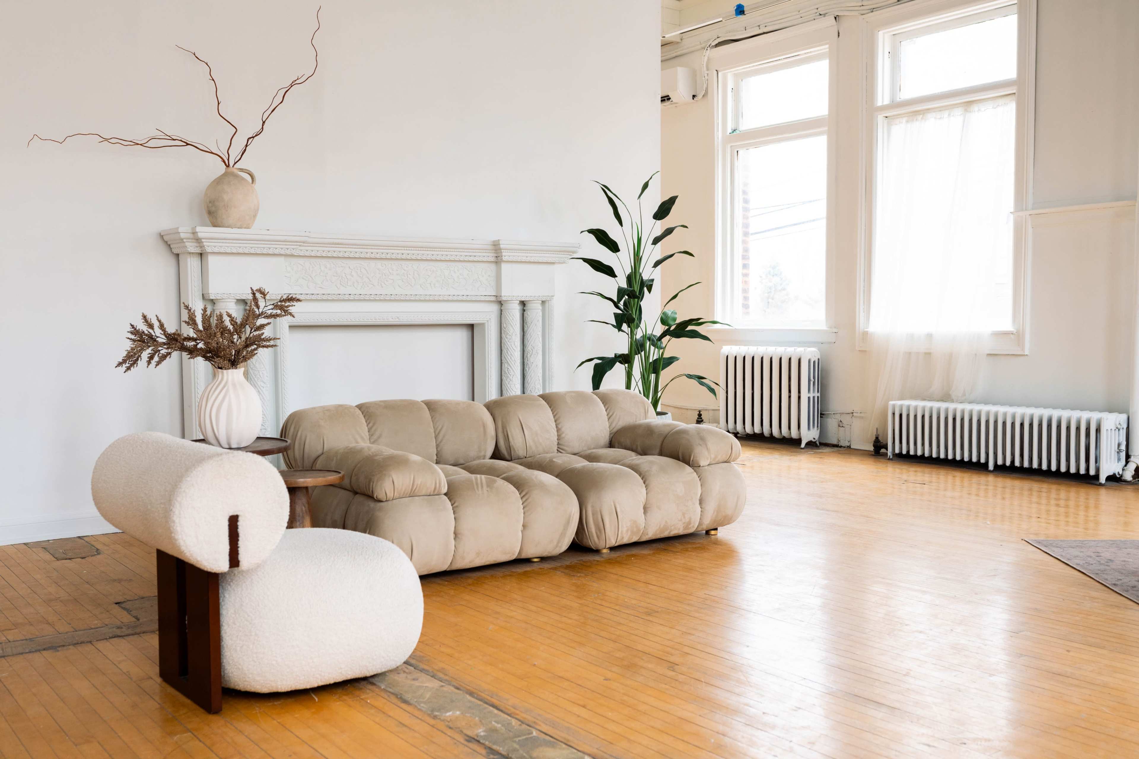 The room features a beige couch and a matching armchair, set against a backdrop of large windows and a decorative fireplace.