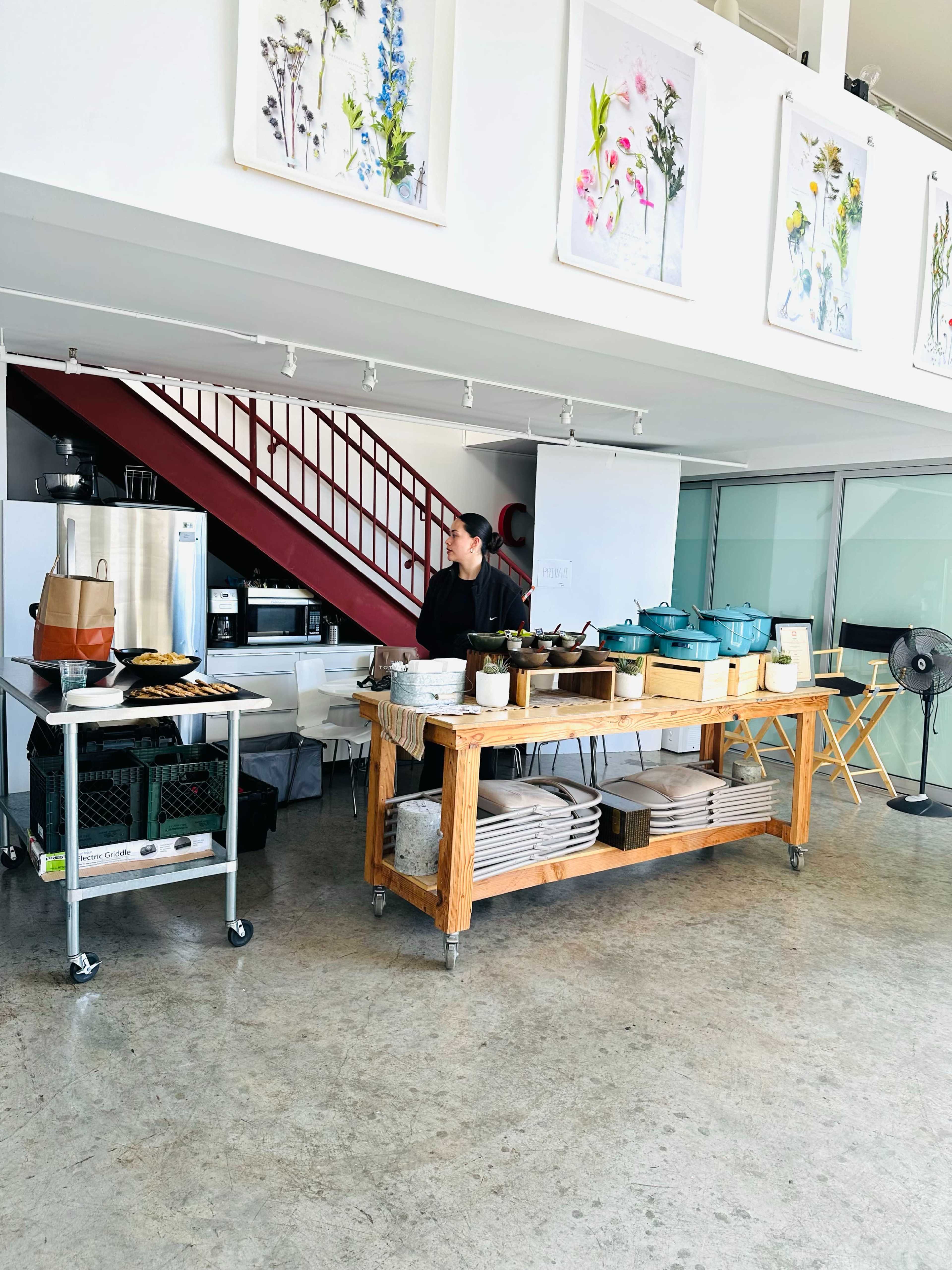A woman stands in a modern kitchen space with a wooden work table and various cooking utensils, under framed artworks of flowers.