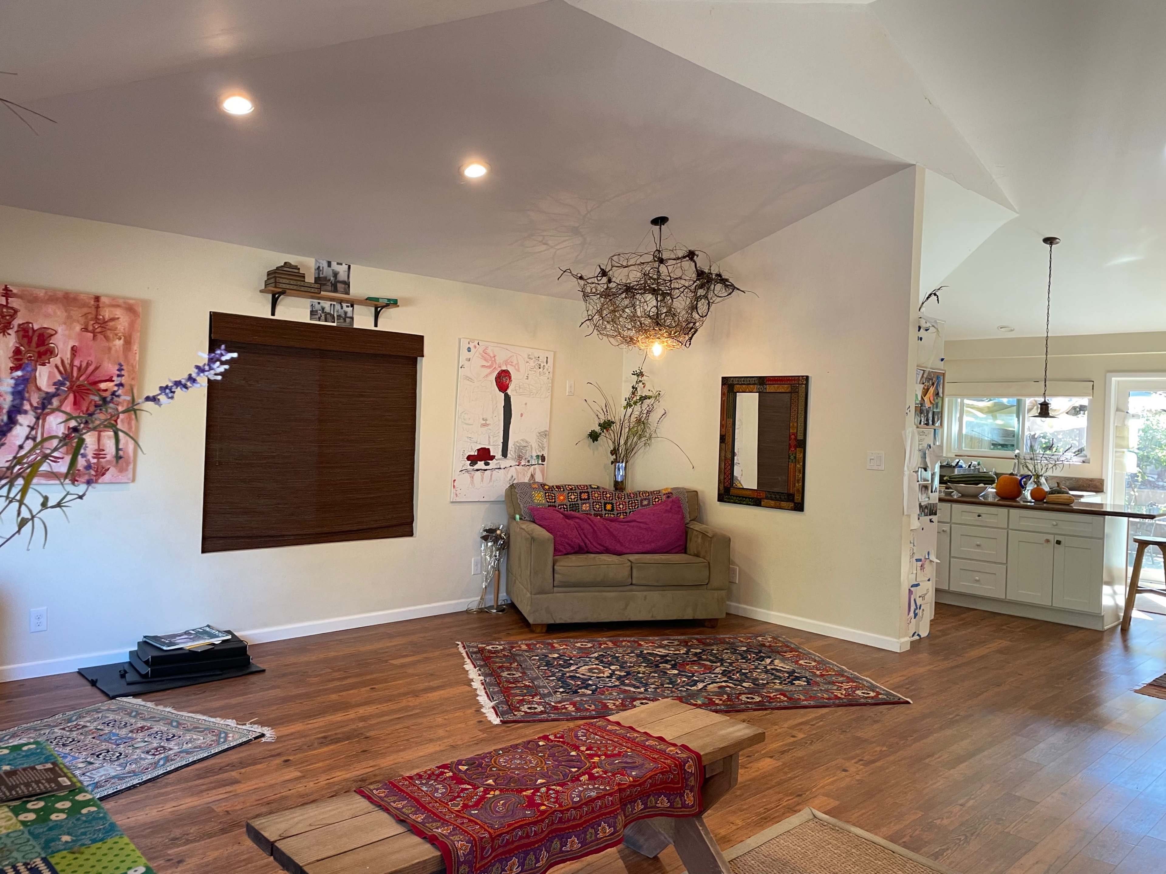 The interior of a home features a cozy seating area with a couch, decorative rugs, and artwork on the walls, leading to a visible kitchen in the background.