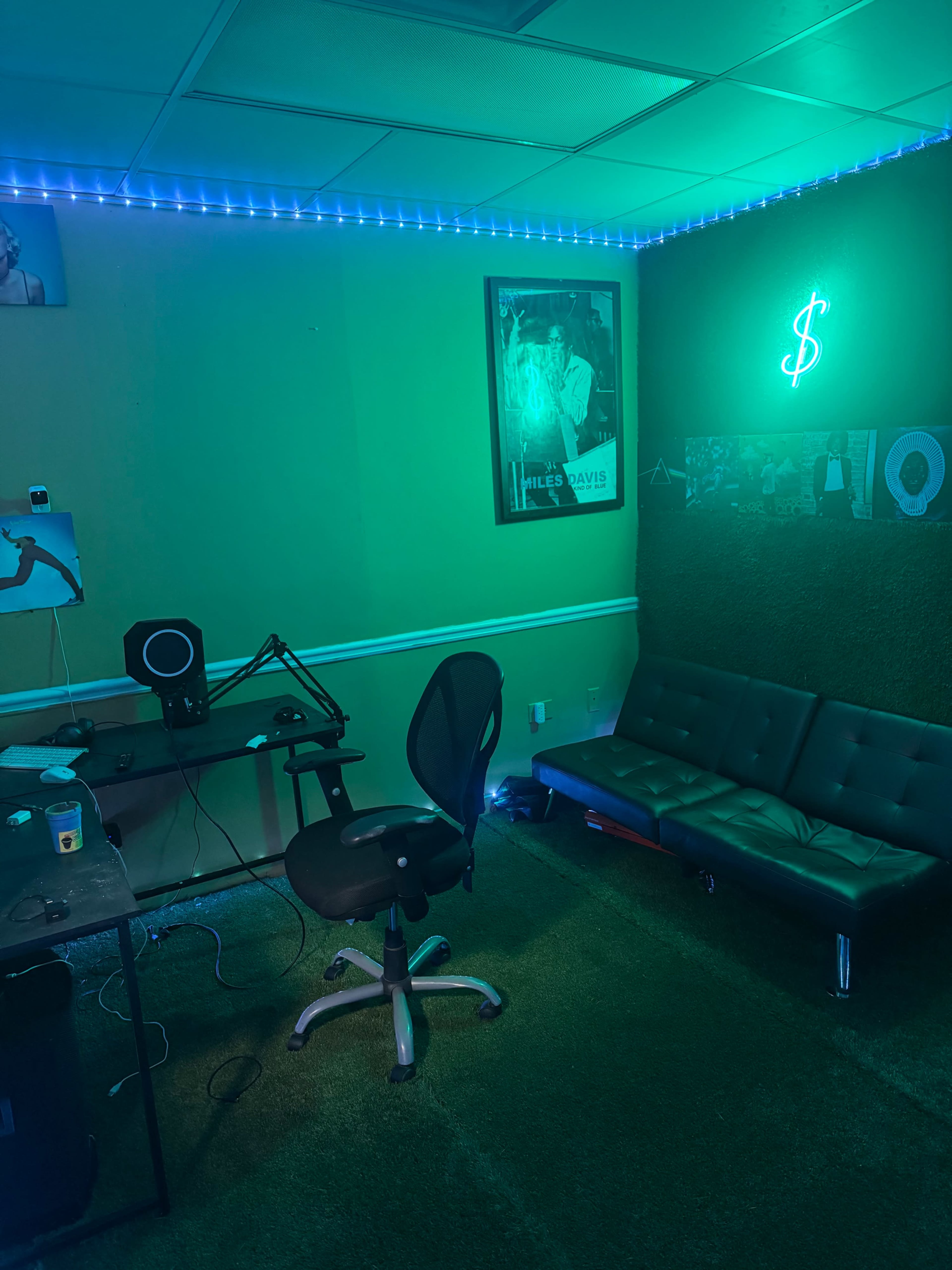 The image shows a dimly lit room with green-tinted walls, a black ergonomic chair at a desk with audio equipment, and a futon against the wall, illuminated by neon lights.