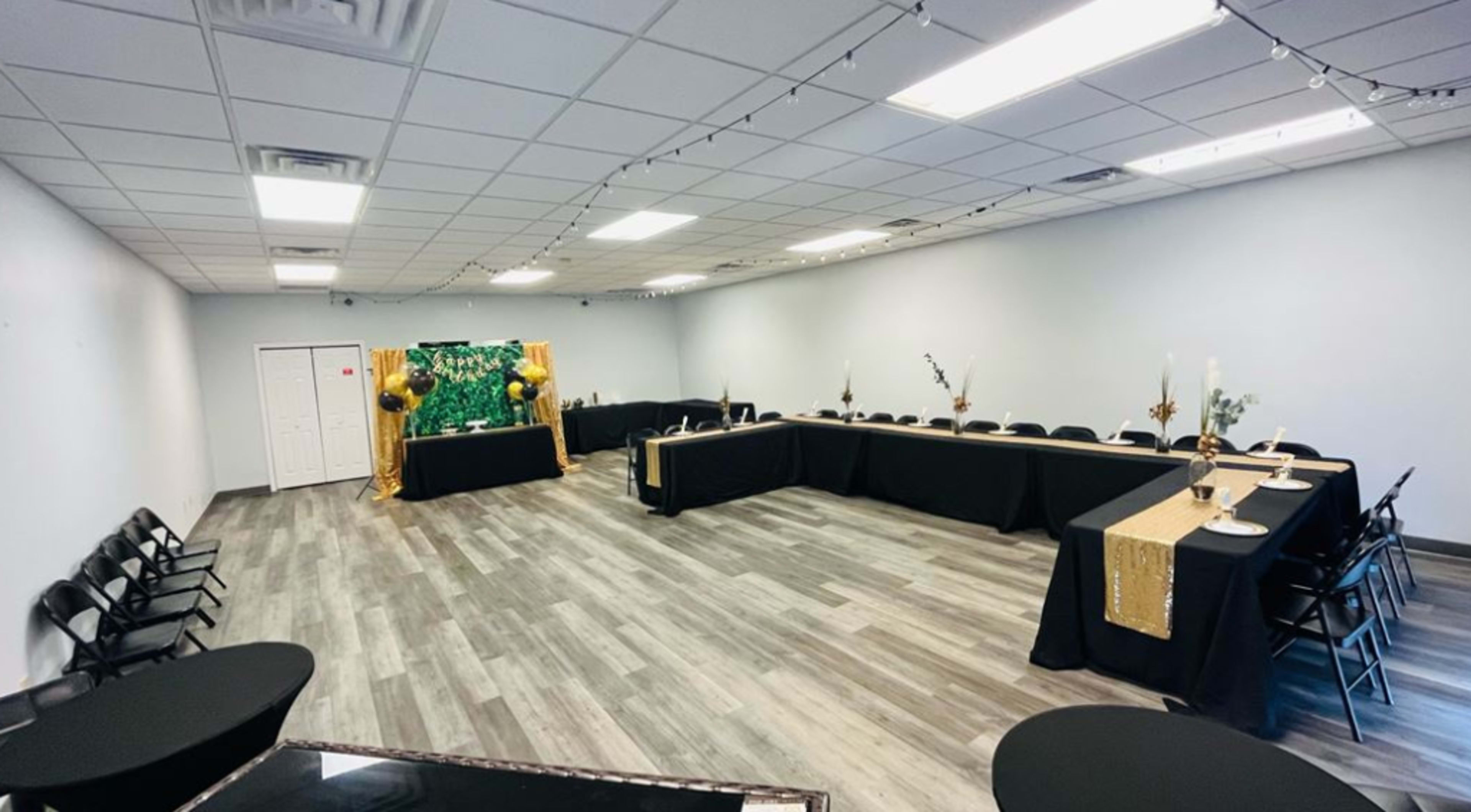 A large, empty event space with multiple tables covered in black cloth, arranged in an open layout, and decorated with gold accents and a green backdrop.