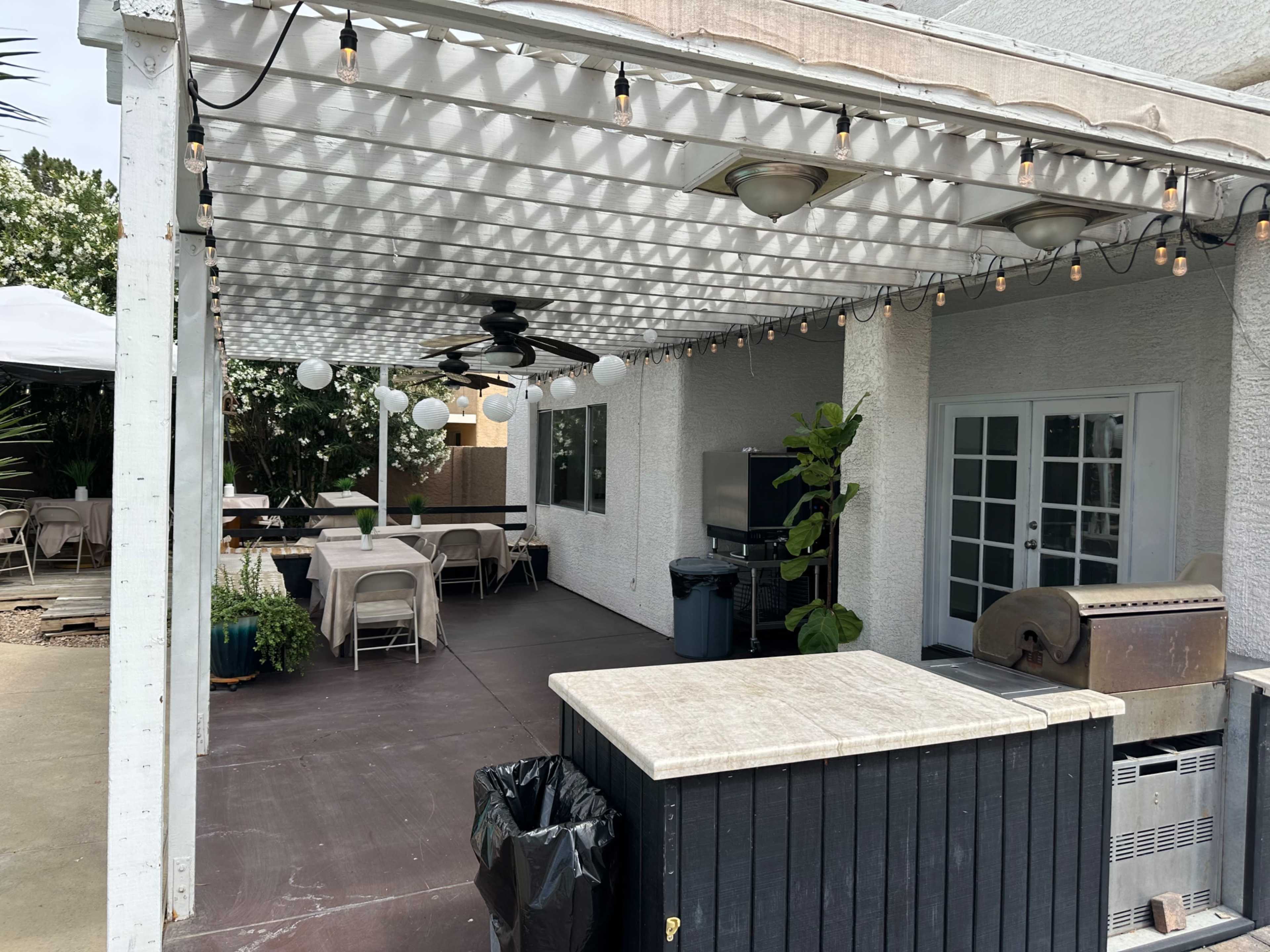 The image shows a covered patio area with string lights, chairs, a fan, and an outdoor table setup.