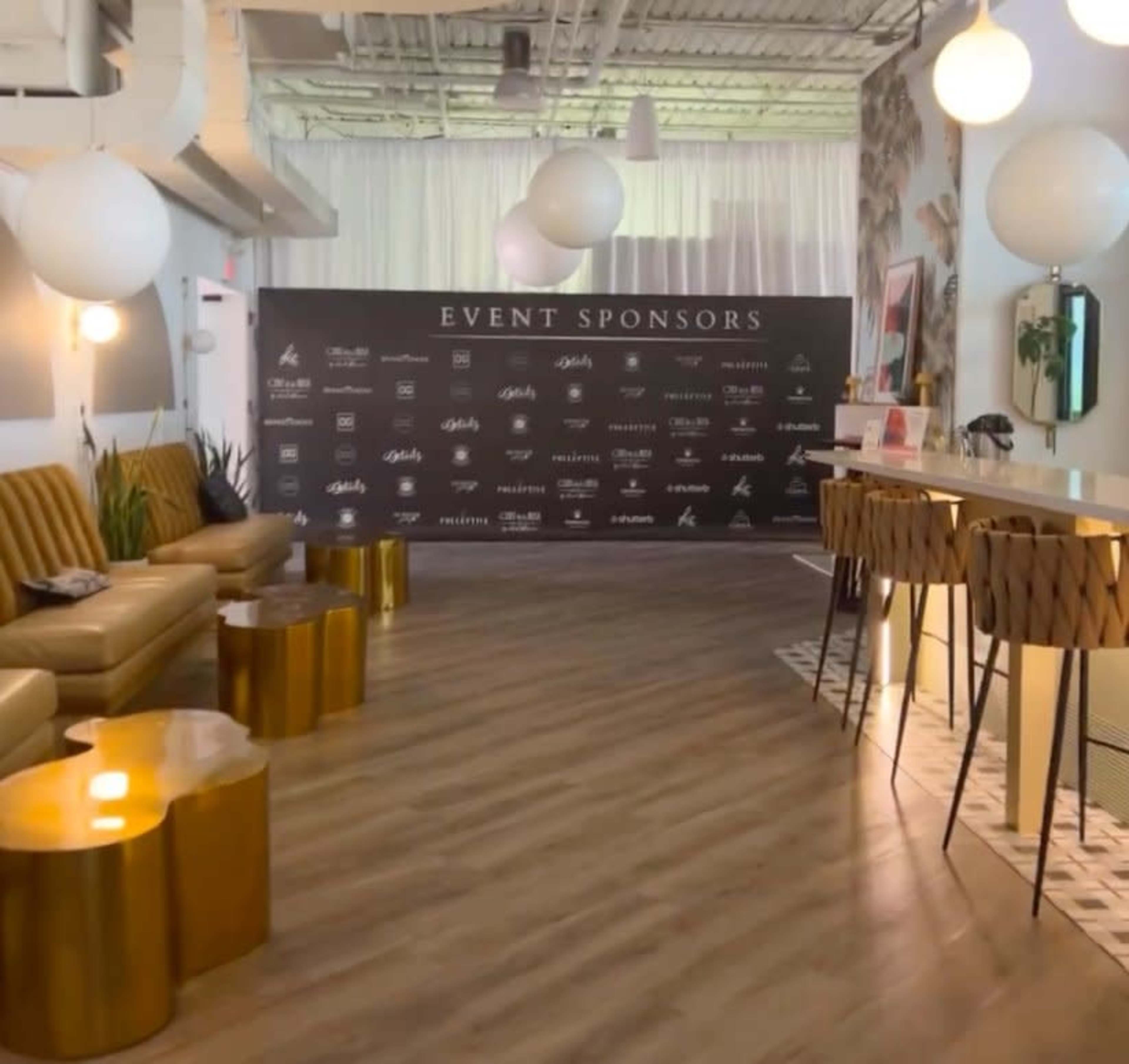 A modern event space with a long brownie leather sofa, round gold accent tables, a bar area, and a backdrop displaying event sponsors.
