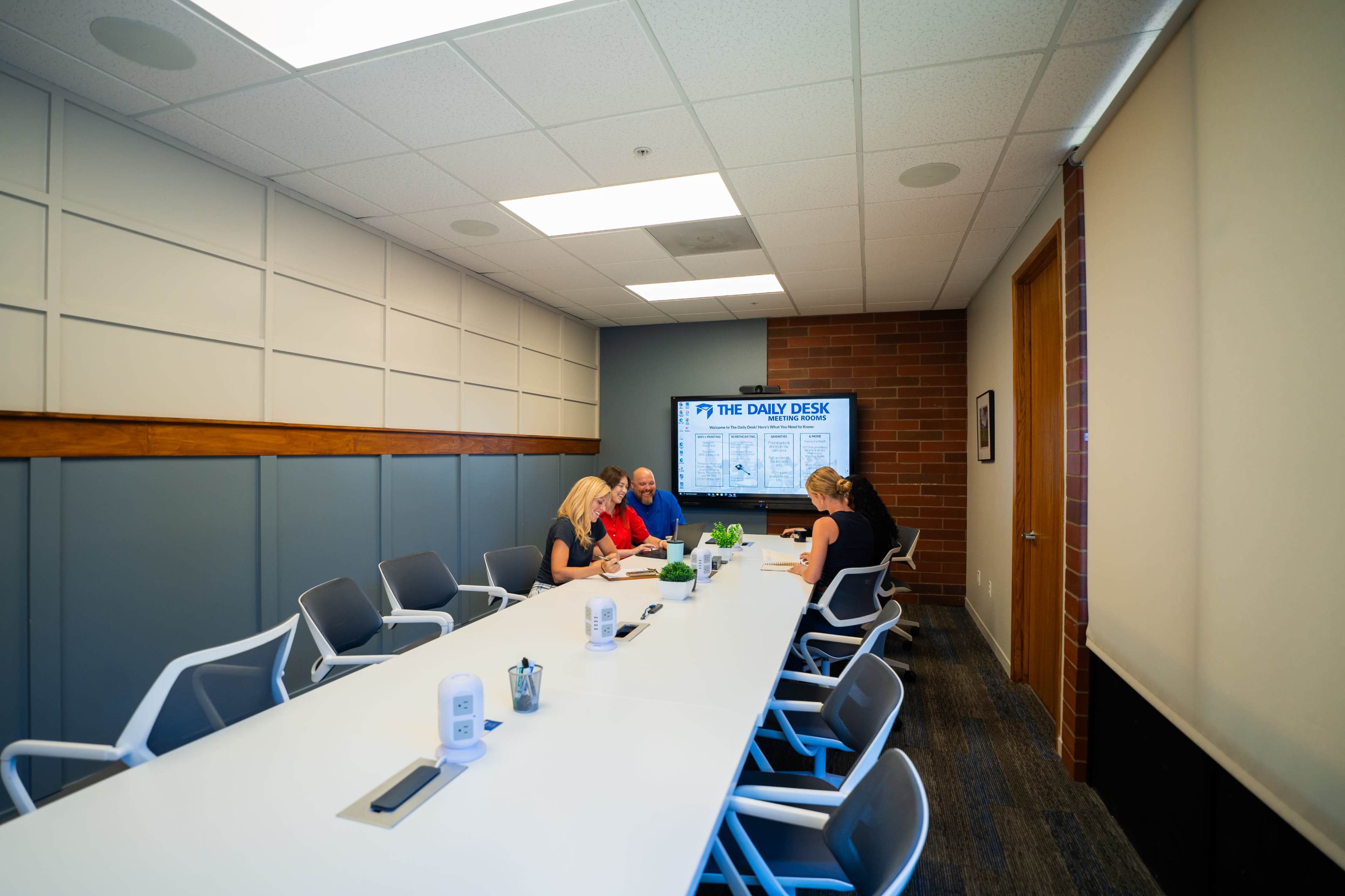 A group of three people is seated around a long, white conference table in a well-lit meeting room, with a presentation screen displaying "THE DAILY DESK" on the wall.
