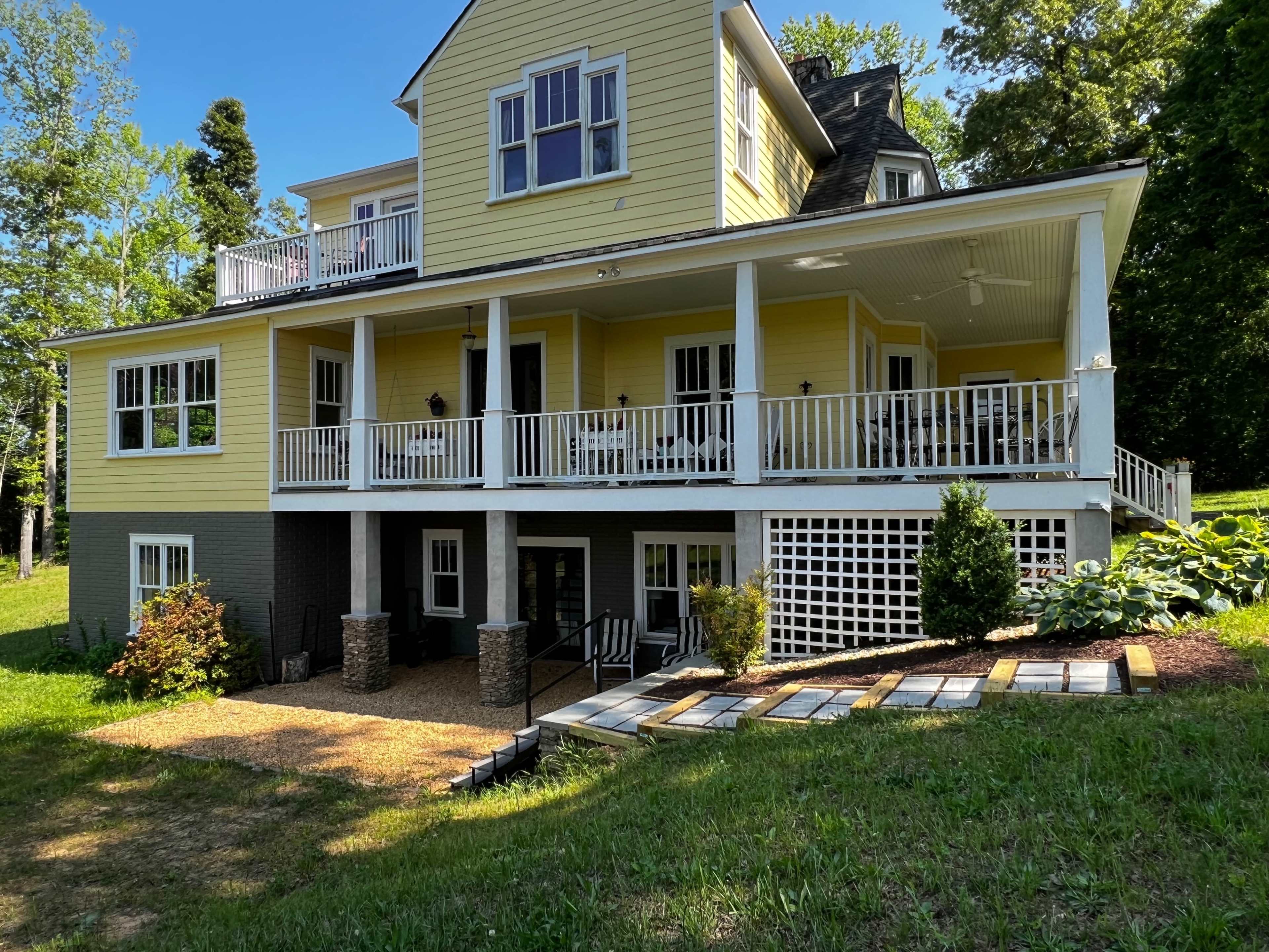 A large yellow house with multiple stories and a covered porch, situated on a grassy slope surrounded by trees.