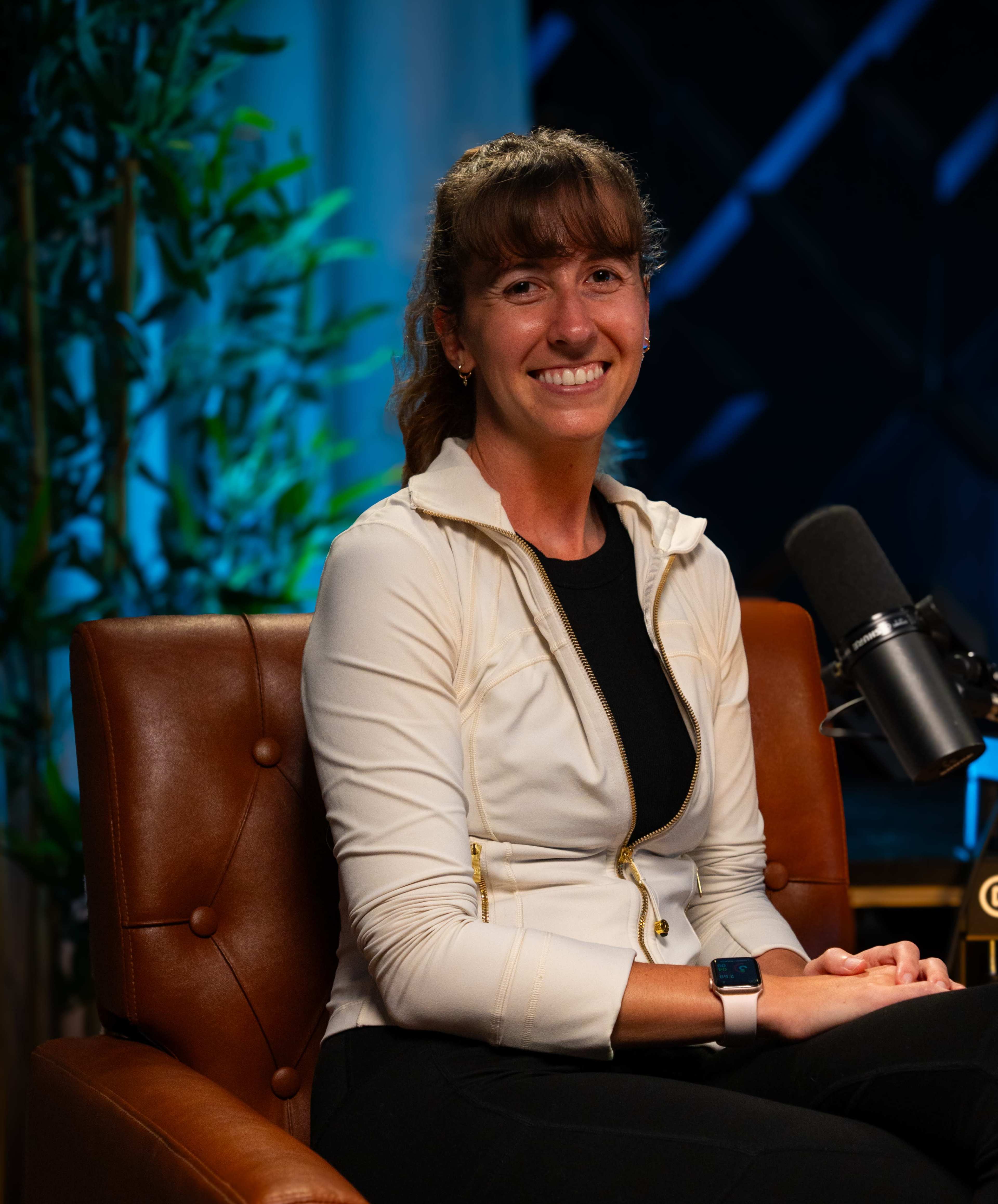 A woman sits on a brown leather chair, smiling at the camera in a well-lit studio setting.
