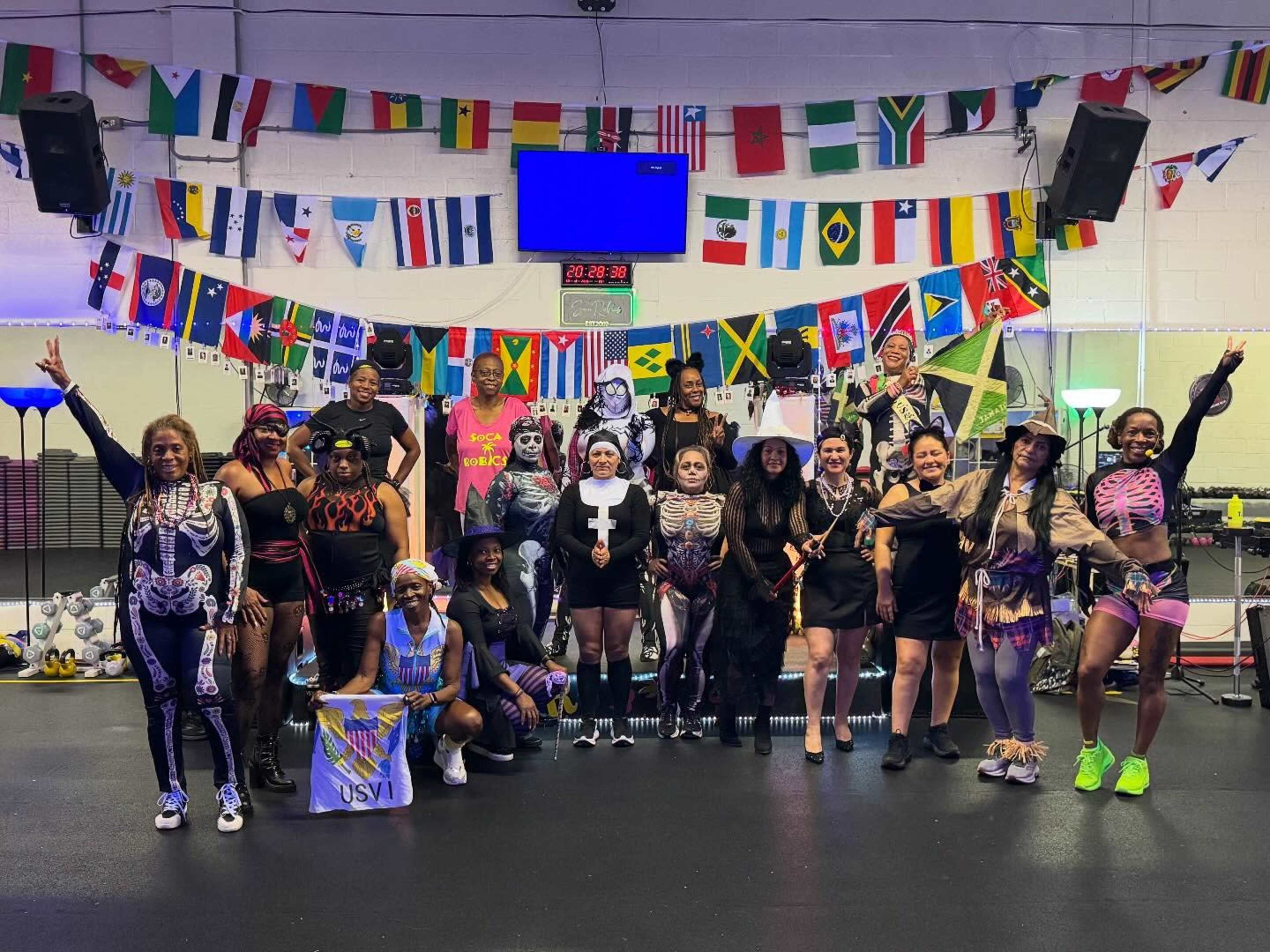 A group of people dressed in creative costumes poses for a photo in a brightly decorated gym, surrounded by flags from various countries.