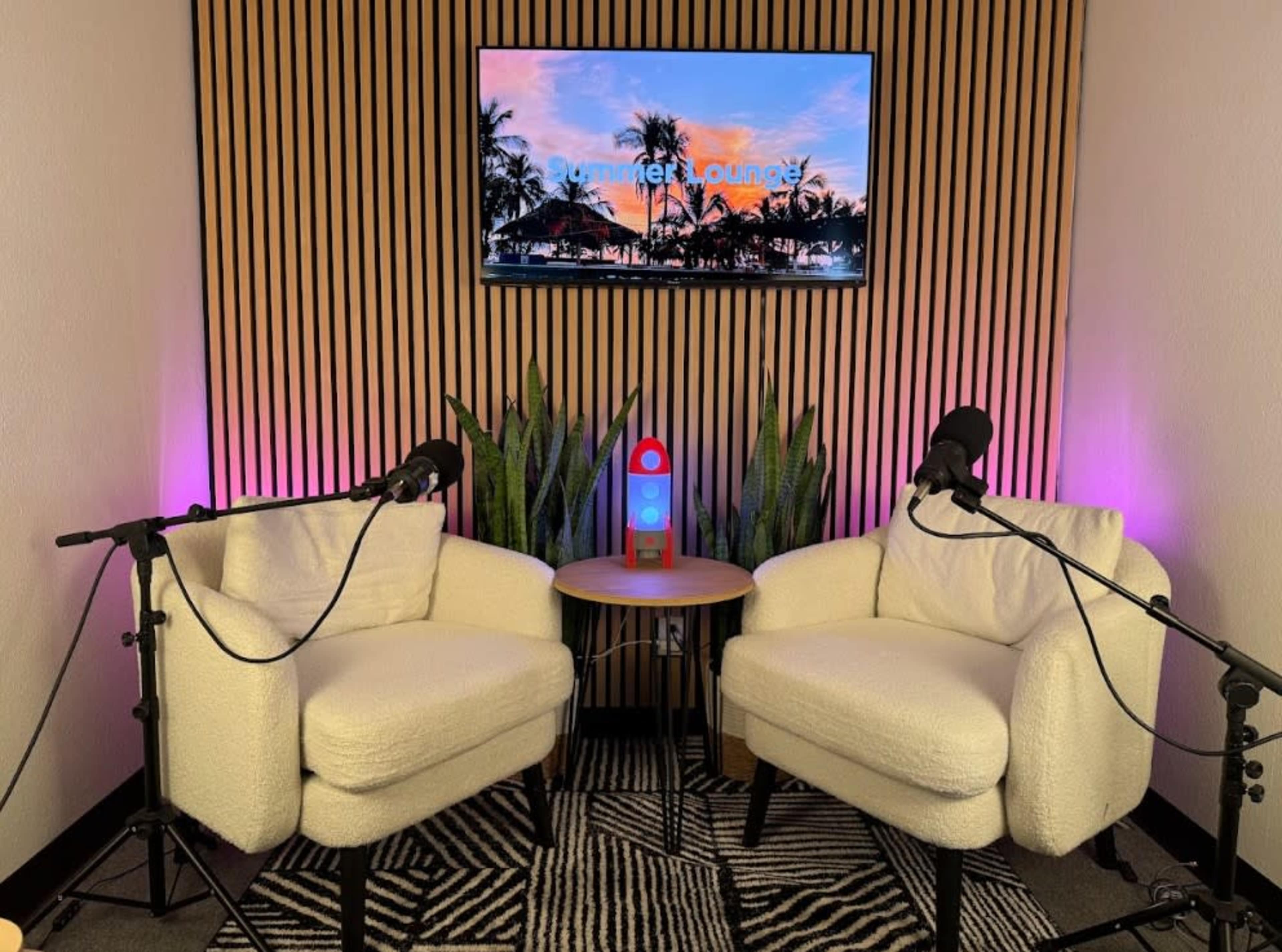 The image shows a cozy audio recording space with two upholstered chairs, a small side table, and a television screen displaying a sunset scene.