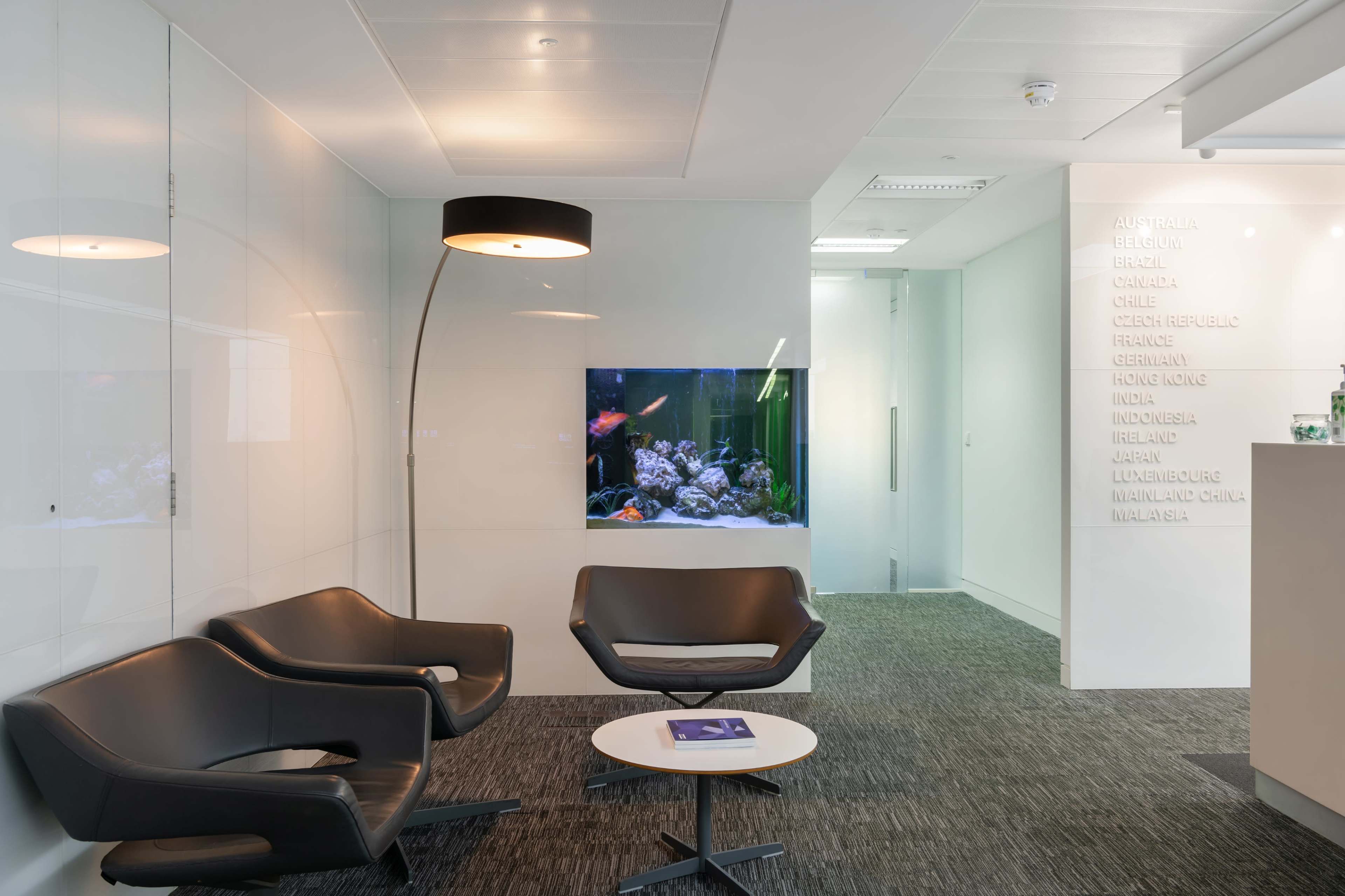 The image shows a modern office waiting area with two black chairs, a round white table, and a fish tank in the background surrounded by a wall with country names inscribed.