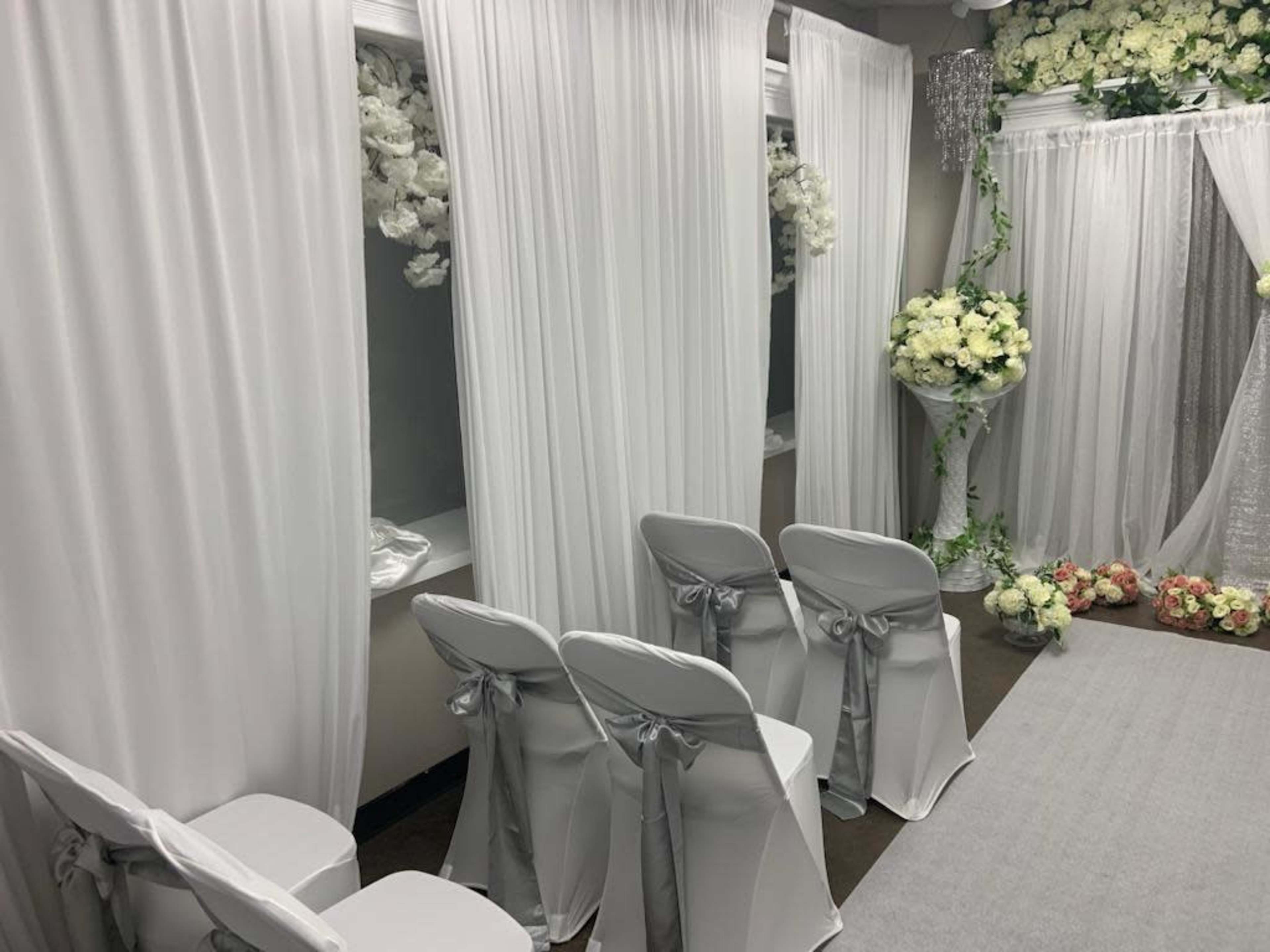 The image shows a wedding ceremony setup featuring white draped fabric, floral arrangements, and chairs with satin bows arranged neatly.
