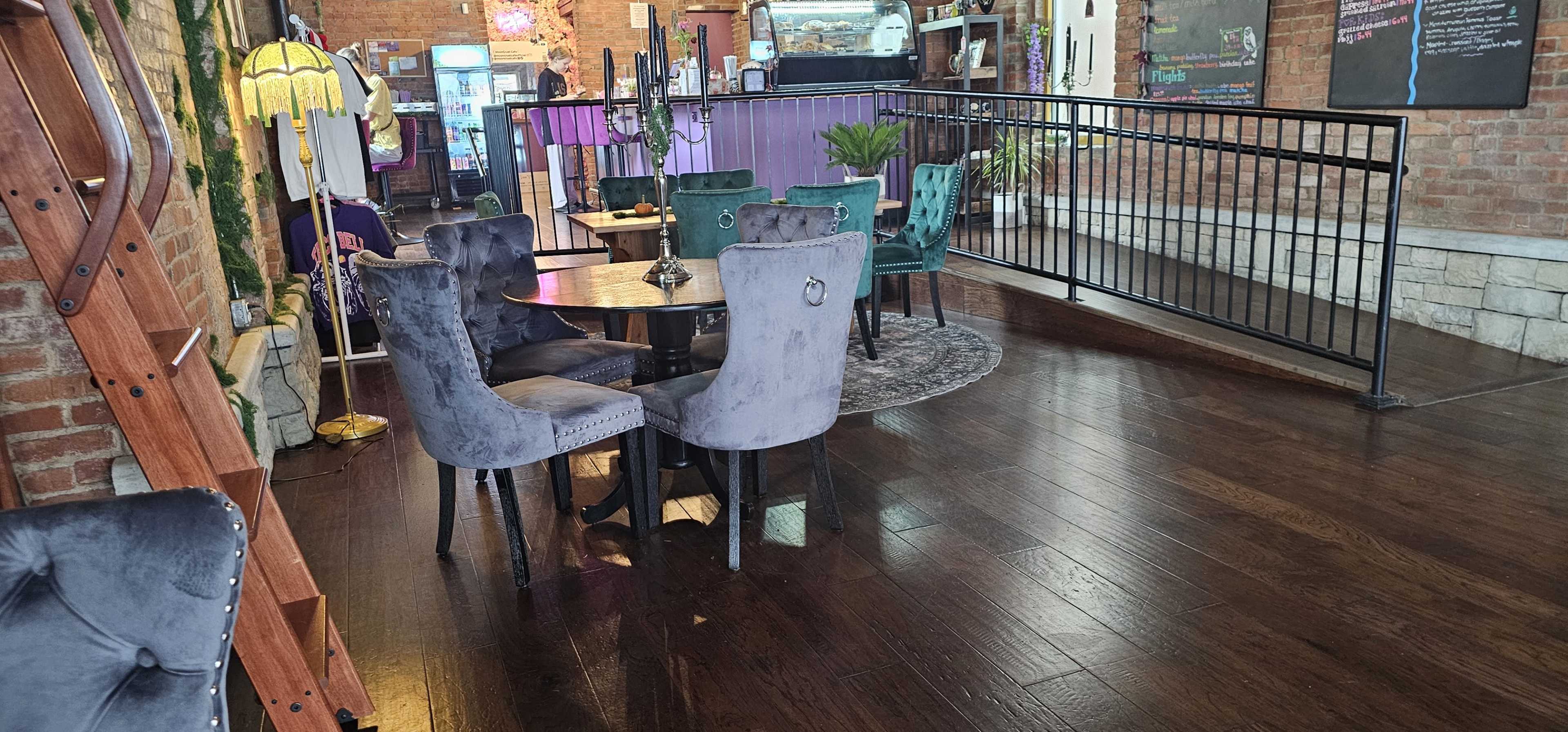 The image shows a cozy café interior with several round tables surrounded by velvet chairs in various colors, set on a polished wooden floor.