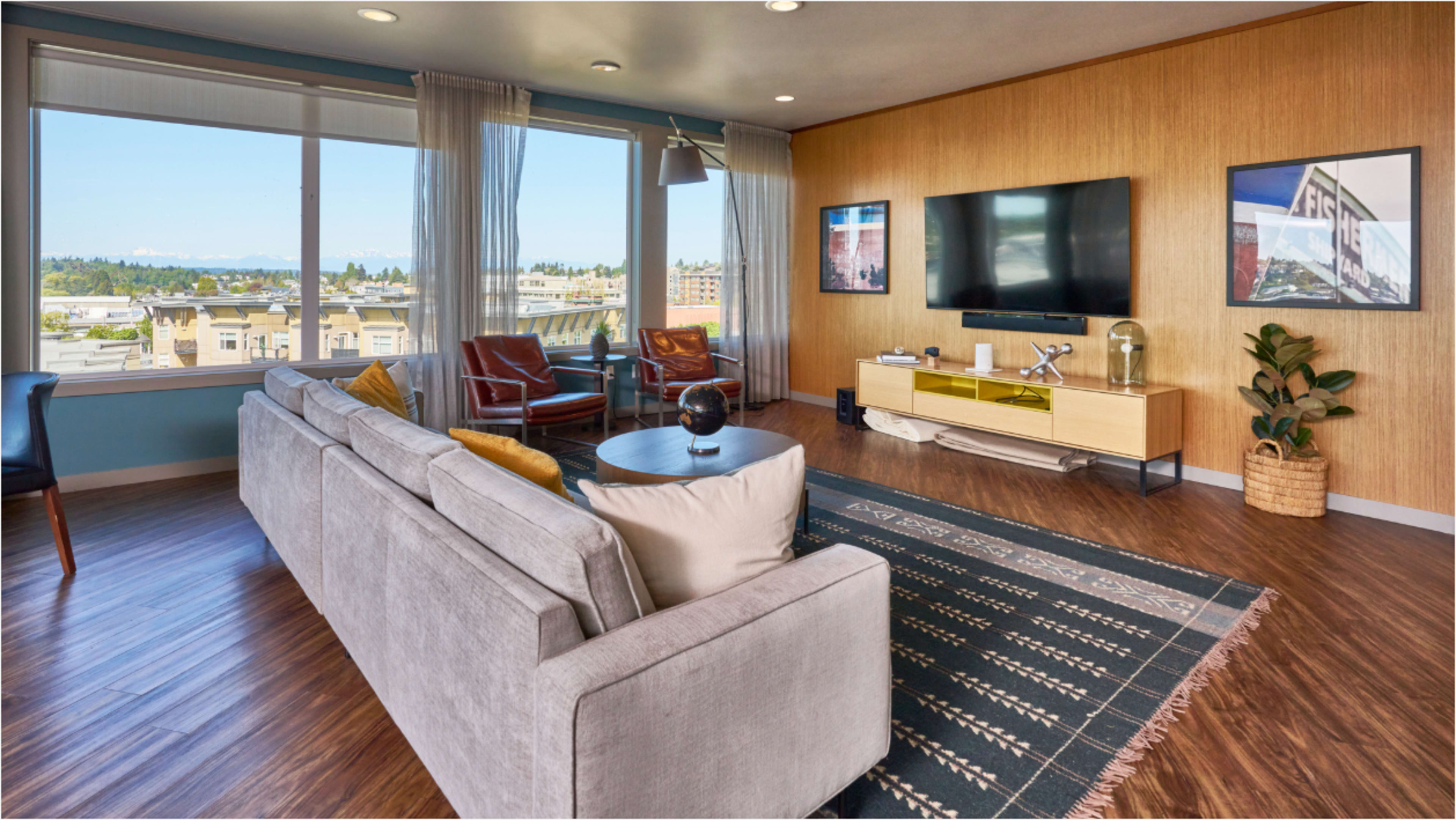 The image shows a modern living room with a large window overlooking a cityscape and mountains, featuring a gray sofa, a coffee table, and a TV on the wall.
