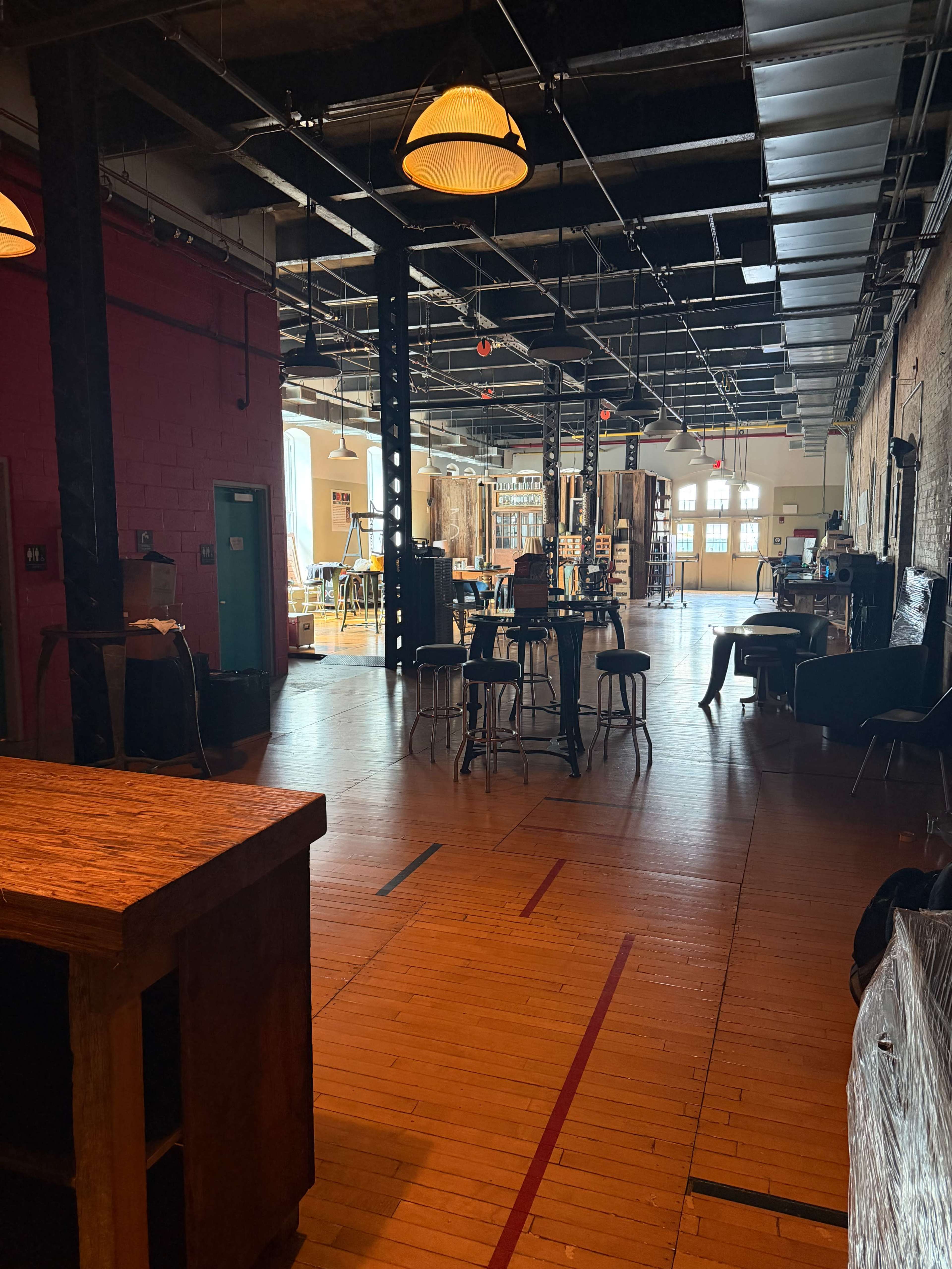 The image shows an open, spacious interior of a rustic venue with wooden flooring, exposed brick walls, and various furniture arrangements including tables and chairs.